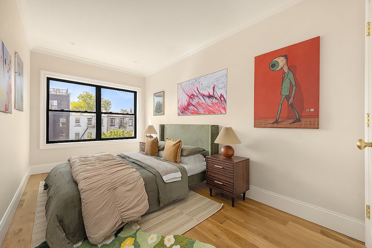 This is a well-lit primary bedroom featuring a queen-sized bed with an olive green headboard and a mix of neutral and earth-toned bedding. A dark wood nightstand with a lamp sits beside the bed, and artwork adorns the walls, adding character to the space. The room has hardwood floors and a large window offering natural light and a view of the outside.
