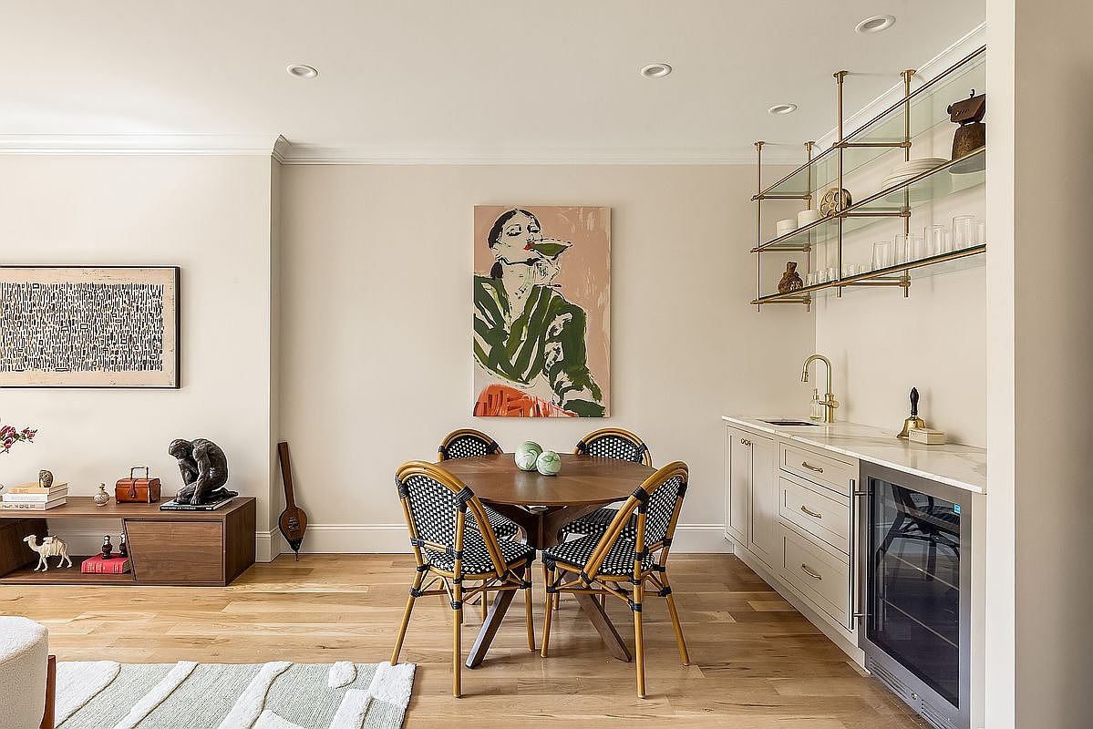 This interior shot showcases a dining area with a round wooden table surrounded by four chairs with woven backs. A large, colorful painting hangs on the wall above the table, and a built-in bar area with glass shelves and a wine fridge is visible to the right. The room features hardwood floors and neutral wall colors, creating a warm and inviting atmosphere.