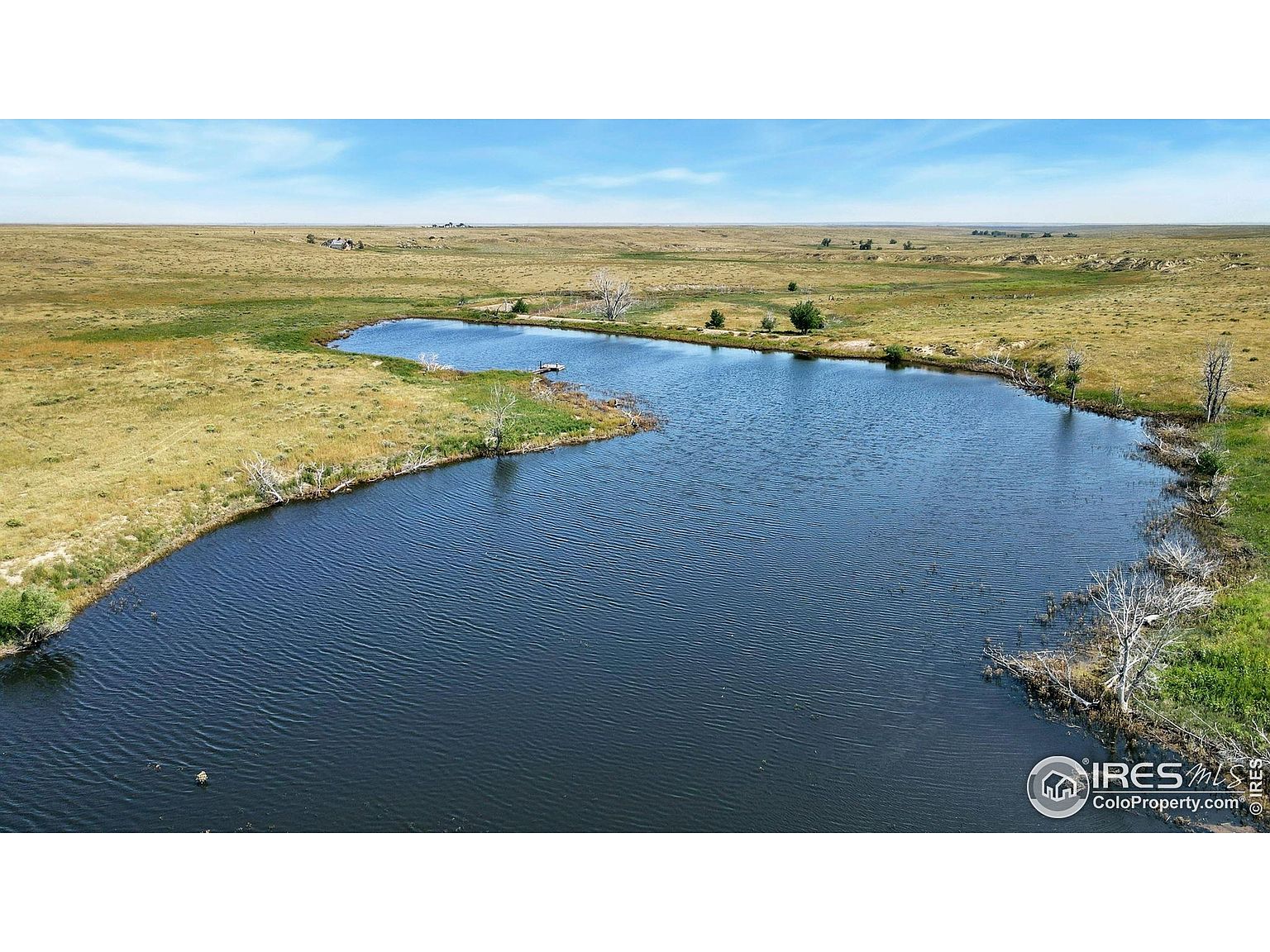 This aerial shot showcases a serene pond surrounded by expansive grasslands under a clear blue sky. The landscape is mostly flat with some undulation, and a few scattered trees dot the shoreline. The image conveys a sense of tranquility and vastness, highlighting the property's natural beauty and potential for outdoor activities.