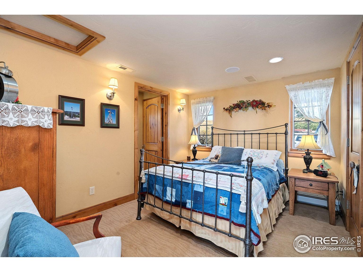 This is a cozy guest bedroom featuring a wrought iron bed with a blue patchwork quilt, wooden nightstand, and warm yellow walls. The room is illuminated by bedside lamps and wall sconces, creating a welcoming atmosphere. The decor includes framed artwork and lace curtains, adding to the room's charm.