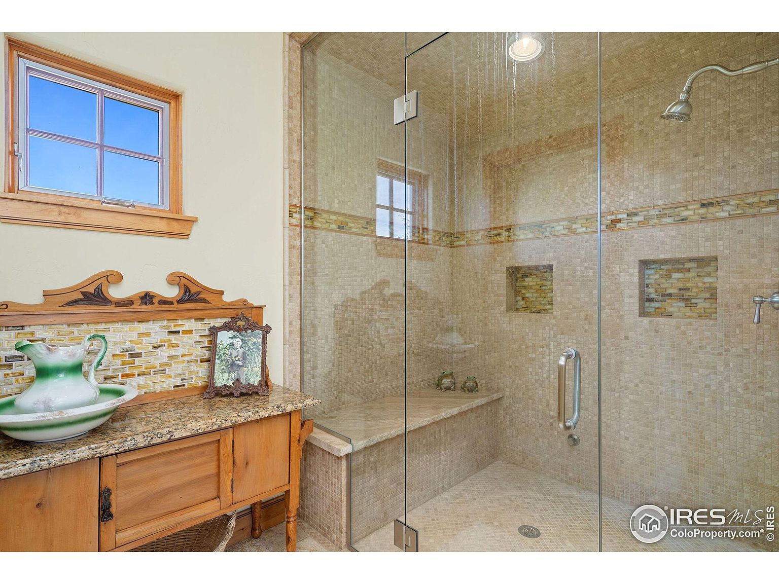 This is a well-appointed primary bathroom featuring a glass-enclosed shower with mosaic tile walls and a built-in bench. A custom vanity with a granite countertop and decorative accents adds character to the space. The overall impression is luxurious and spa-like, with attention to detail in the tile work and fixtures.
