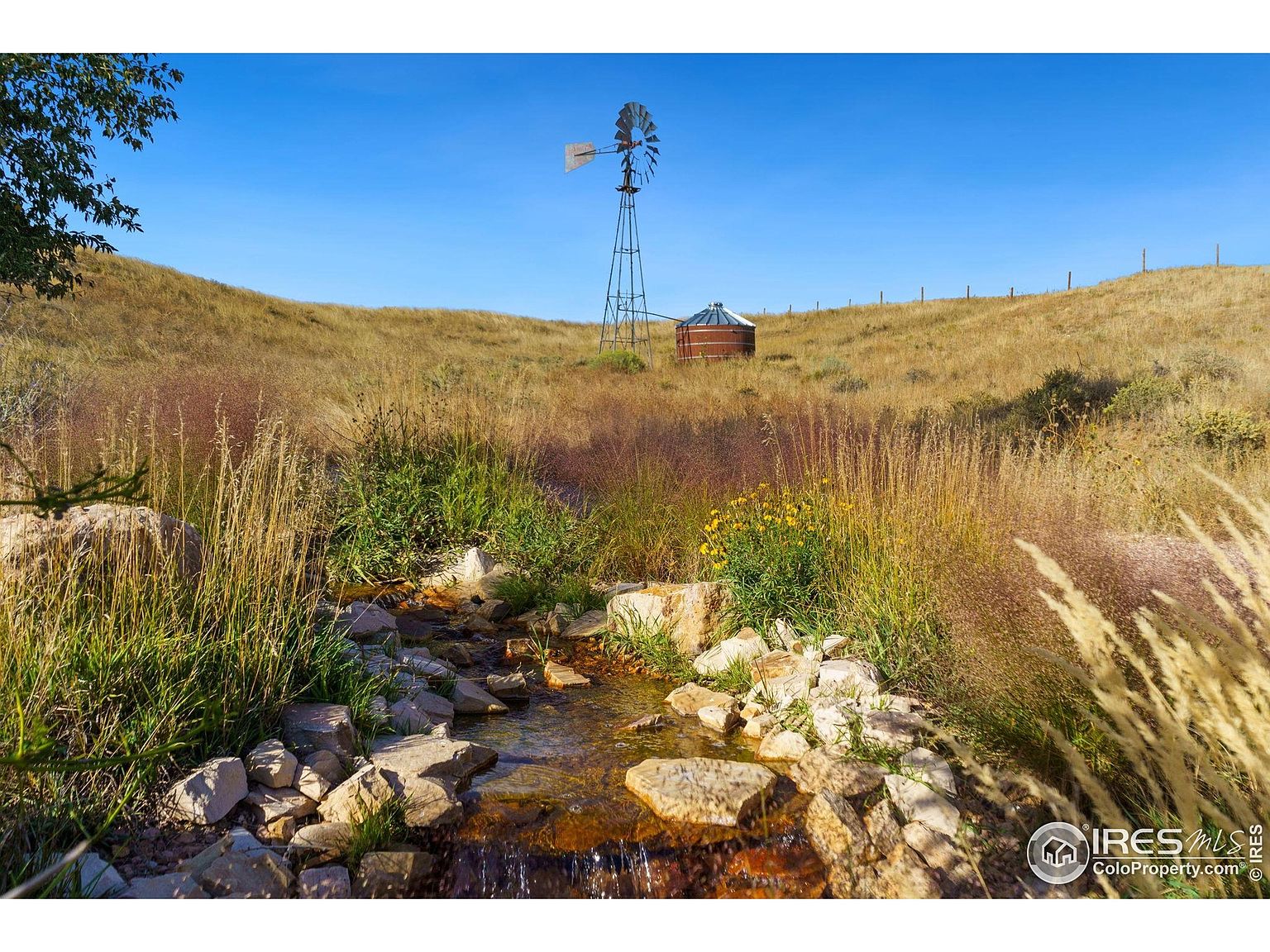 This picturesque yard features a small, rocky stream flowing through lush vegetation, creating a serene and natural landscape. In the background, a classic windmill and a rustic water tank add a touch of rural charm, set against a backdrop of rolling hills and a clear blue sky. The scene evokes a sense of tranquility and natural beauty, highlighting the property's unique outdoor features.