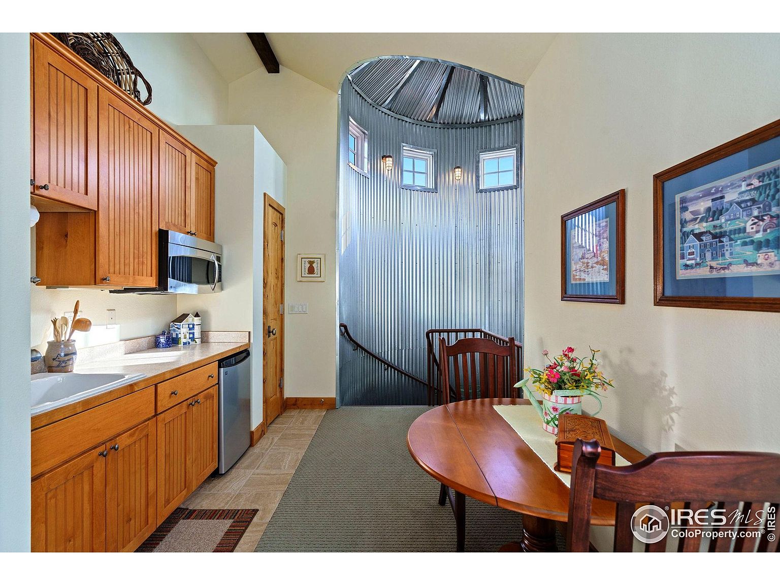 This interior shot showcases a dining area adjacent to a kitchen space. The dining area features a round wooden table with chairs, complemented by artwork on the walls. A unique architectural element is the cylindrical metal structure with windows, adding character to the space. The room is well-lit and appears cozy.