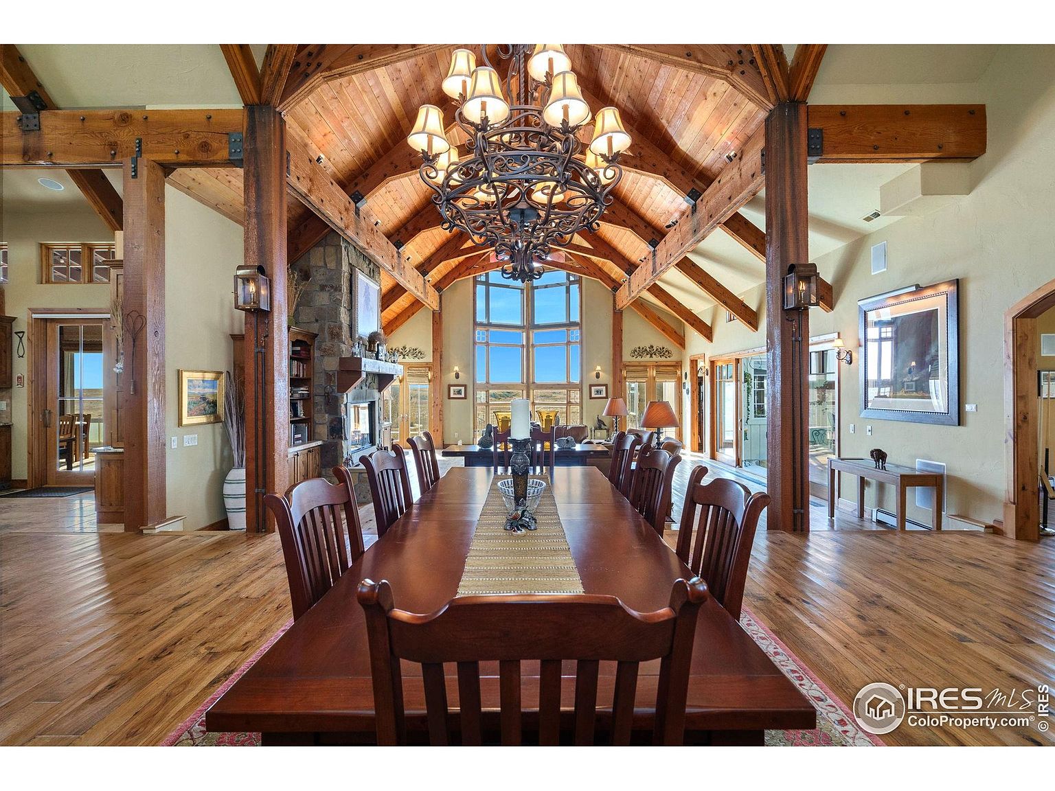 This is an interior shot of a grand dining room featuring a long, dark wood dining table with numerous chairs, suggesting ample seating for large gatherings. The room boasts a high, vaulted ceiling with exposed wooden beams and a large, ornate chandelier. Large windows at the far end of the room provide natural light and a view to the outside, while wooden pillars add architectural interest and divide the space.