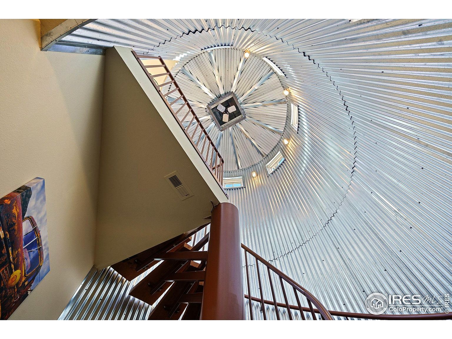 This image showcases a unique interior featuring a spiral staircase within a cylindrical structure, possibly a silo conversion. The walls are lined with corrugated metal, creating a distinctive architectural detail. A wooden staircase with metal railings ascends, and a square skylight at the top provides natural light, enhancing the space's character.