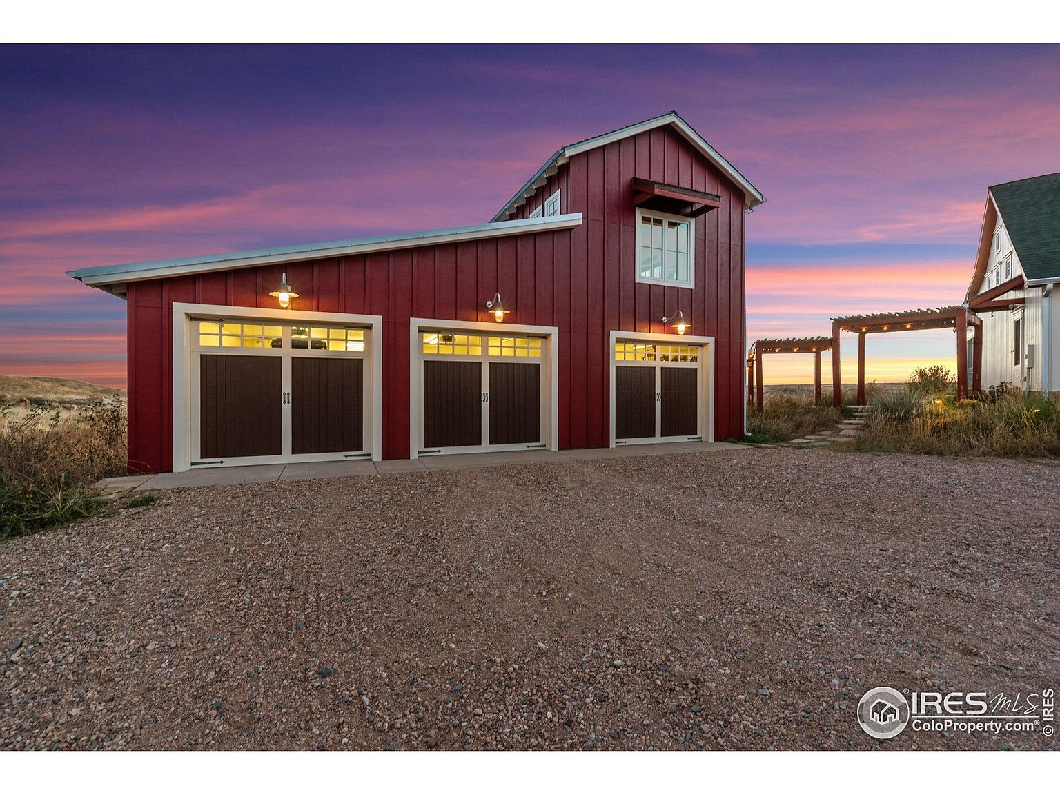 This image showcases a charming red barn-style garage with three bays, each featuring dark brown doors with white trim. The garage is well-lit with exterior lights above each door, and the gravel driveway leads up to the structure. The setting includes a glimpse of a pergola and another building, all under a vibrant sunset sky, creating a picturesque and inviting scene.