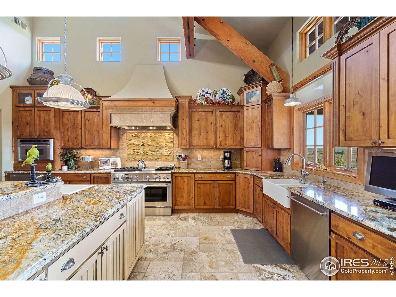 This is a well-lit kitchen featuring custom wood cabinetry, granite countertops, and stainless steel appliances. The kitchen includes a large island with a white base and granite top, a farmhouse sink, and a decorative tile backsplash. The space is accented by a wooden beam and multiple windows providing natural light.