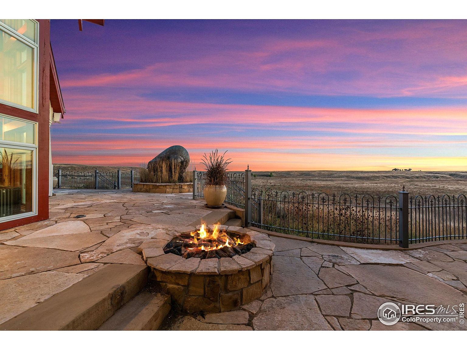 This image showcases a stunning outdoor patio area at sunset. A stone fire pit takes center stage, with flames flickering against the backdrop of a vibrant sky. The patio is constructed from flagstone and features a decorative fence, offering an inviting space for relaxation and entertainment.