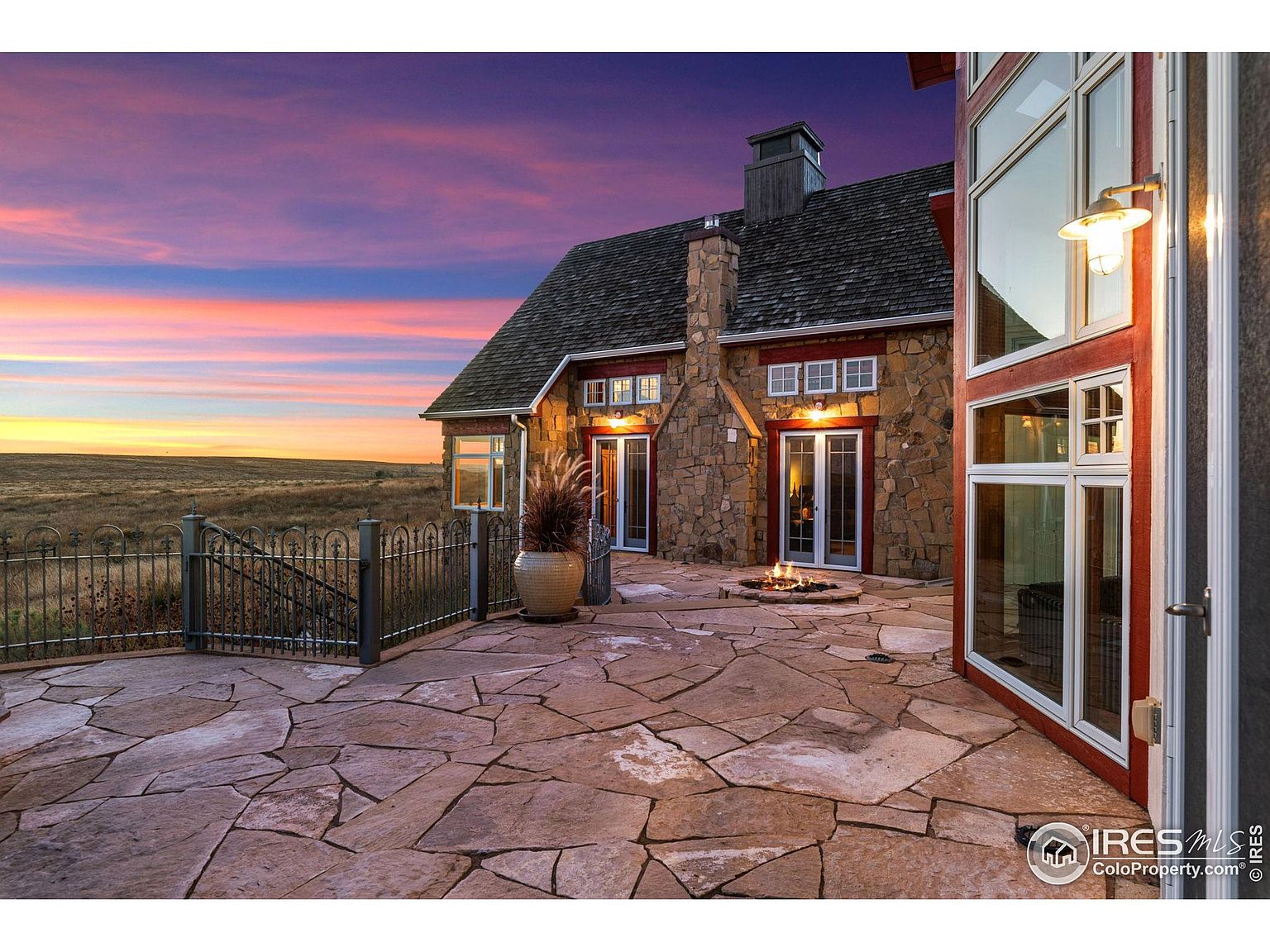 This image showcases the rear exterior of a stone-clad house at dusk, featuring a flagstone patio with a fire pit. The house has a prominent stone chimney and multiple windows, with a wrought-iron fence bordering the property. The sky is painted with vibrant hues of pink and orange, creating a warm and inviting atmosphere.