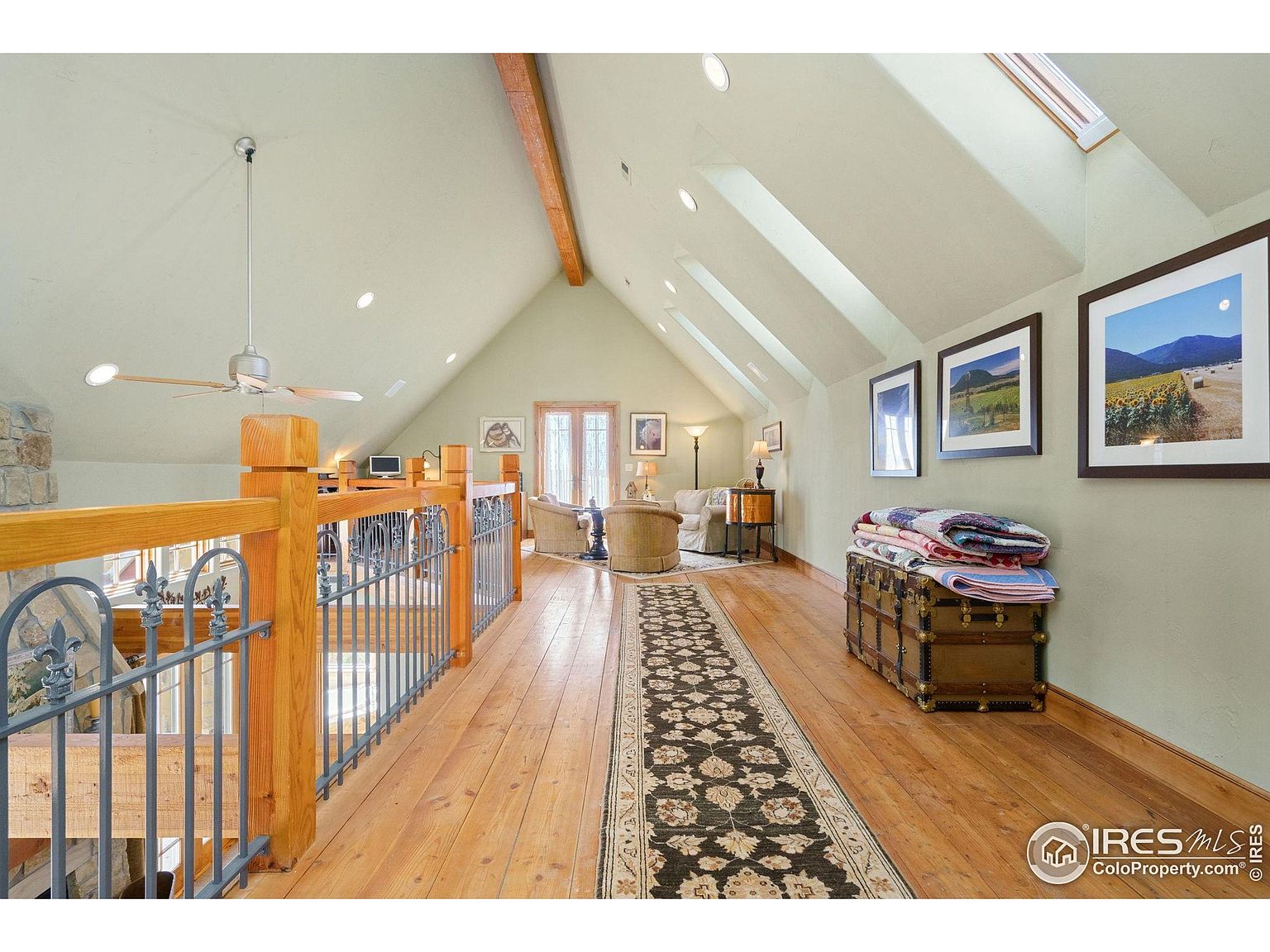 This image showcases an interior hallway or landing area, featuring hardwood floors and a vaulted ceiling with skylights. A wooden railing with metal accents overlooks a lower level, and framed artwork adorns the walls. A vintage trunk with folded blankets adds a touch of character to the space.