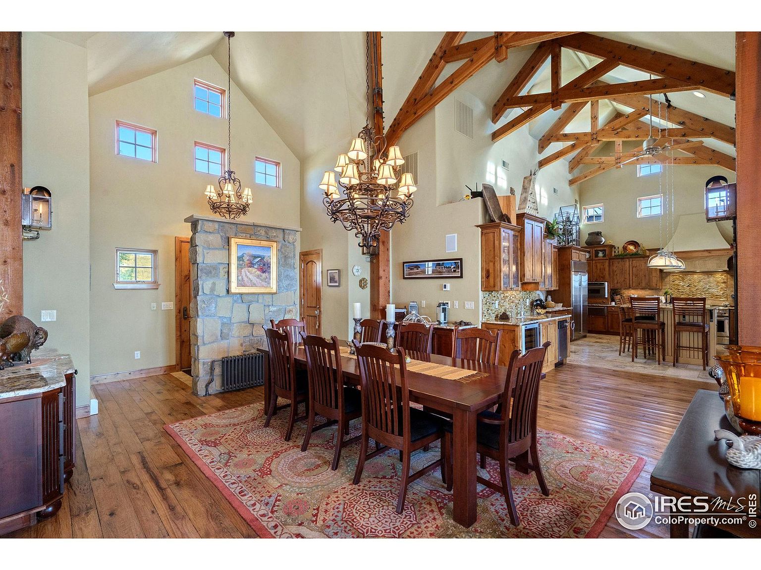 This interior shot showcases a dining room with a large wooden table and chairs set on an ornate rug. The room features a high vaulted ceiling with exposed wooden beams and a decorative chandelier. The dining area seamlessly transitions into an open-concept kitchen, creating a spacious and inviting atmosphere.