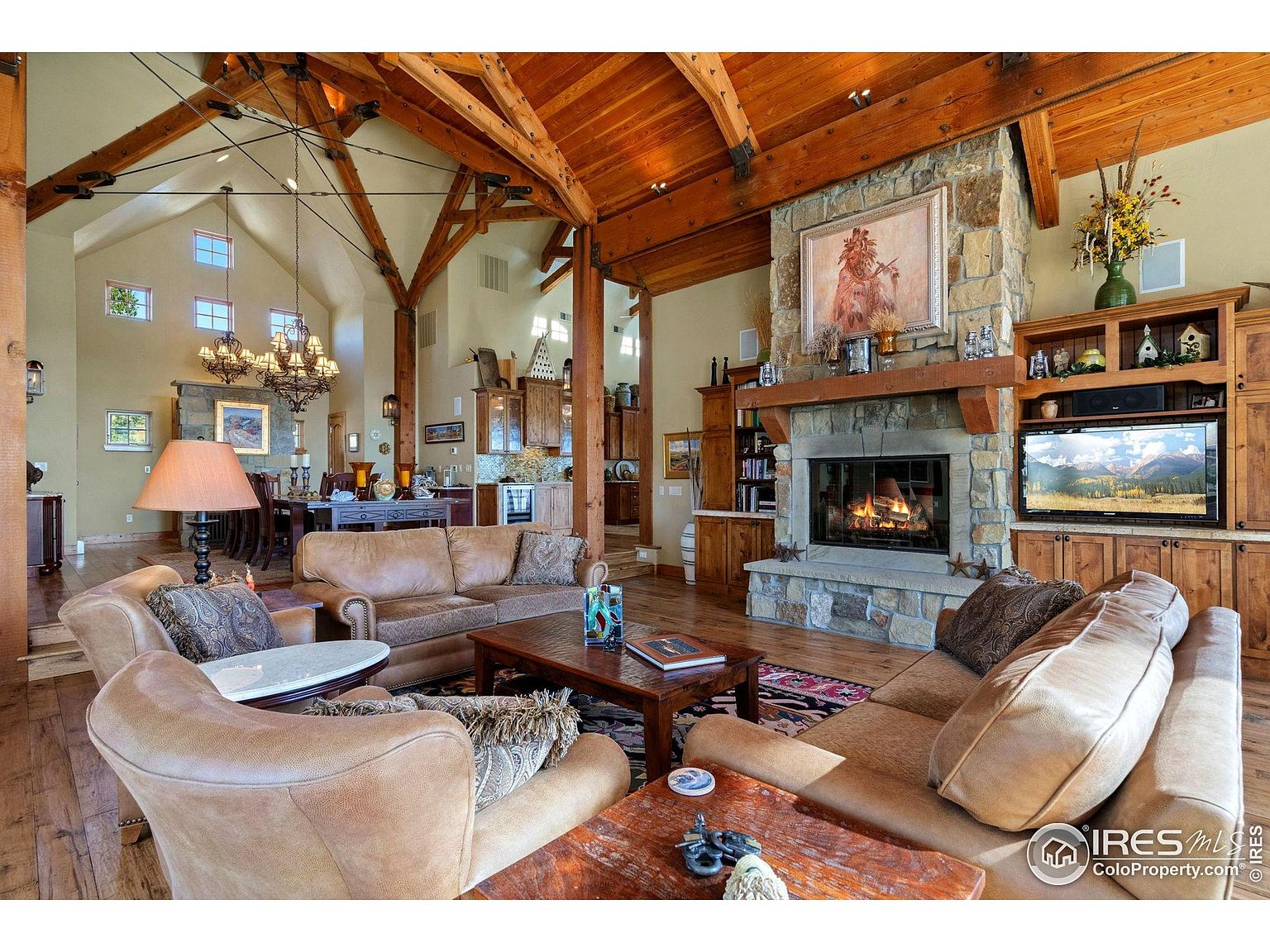 This is a spacious living room featuring a stone fireplace with a wooden mantel and a large painting above it. The room has high, vaulted ceilings with exposed wooden beams, creating a rustic yet elegant atmosphere. Comfortable leather sofas and armchairs are arranged around a coffee table, and a large television is built into a wooden cabinet, suggesting a cozy and inviting space for relaxation and entertainment.