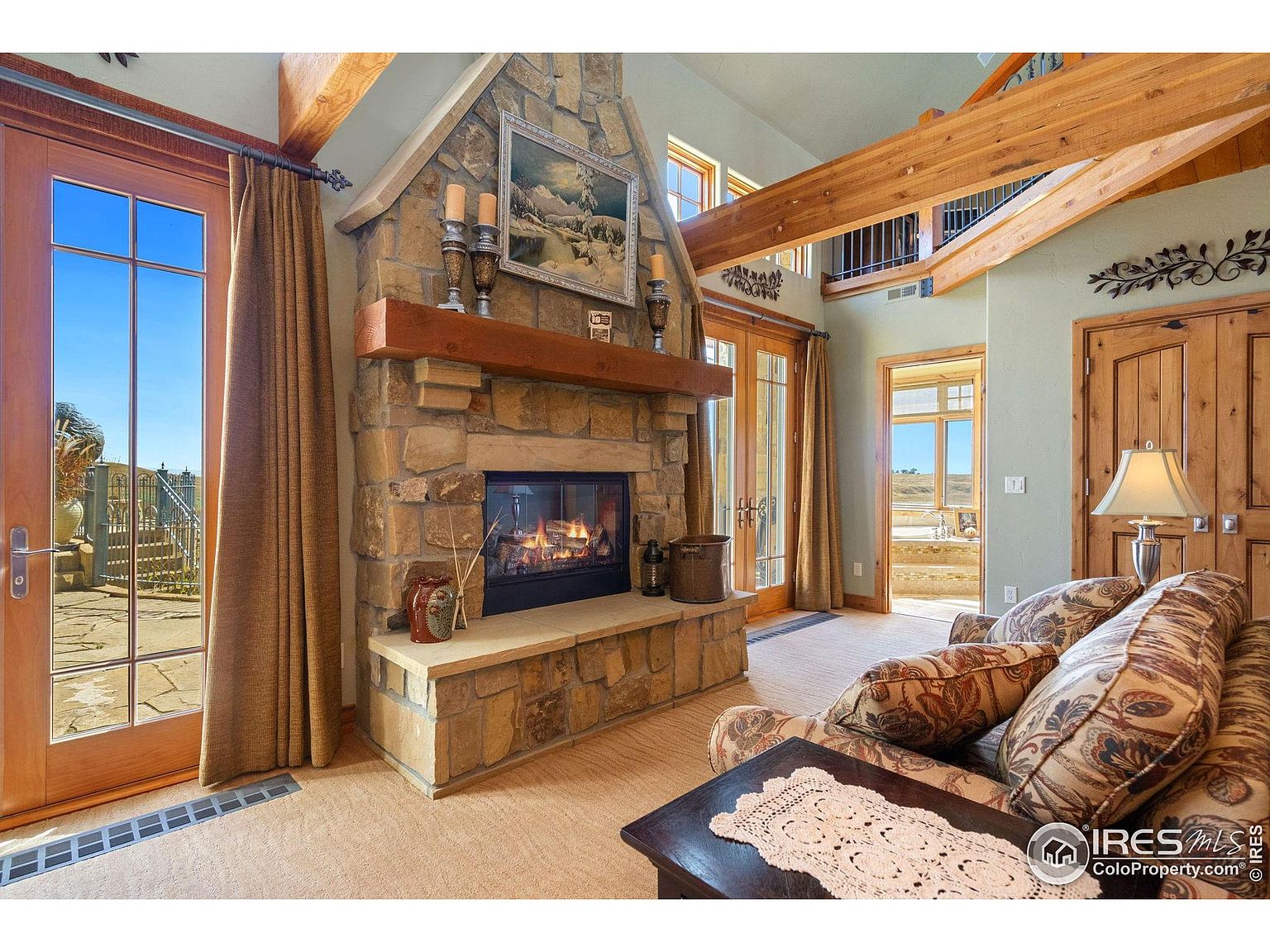 This is an interior shot of a living room featuring a stone fireplace as a focal point. The room has wooden beams on the ceiling and large windows/doors that let in natural light. The furniture includes a patterned sofa and a dark wood coffee table with a lace runner, creating a cozy and inviting atmosphere.