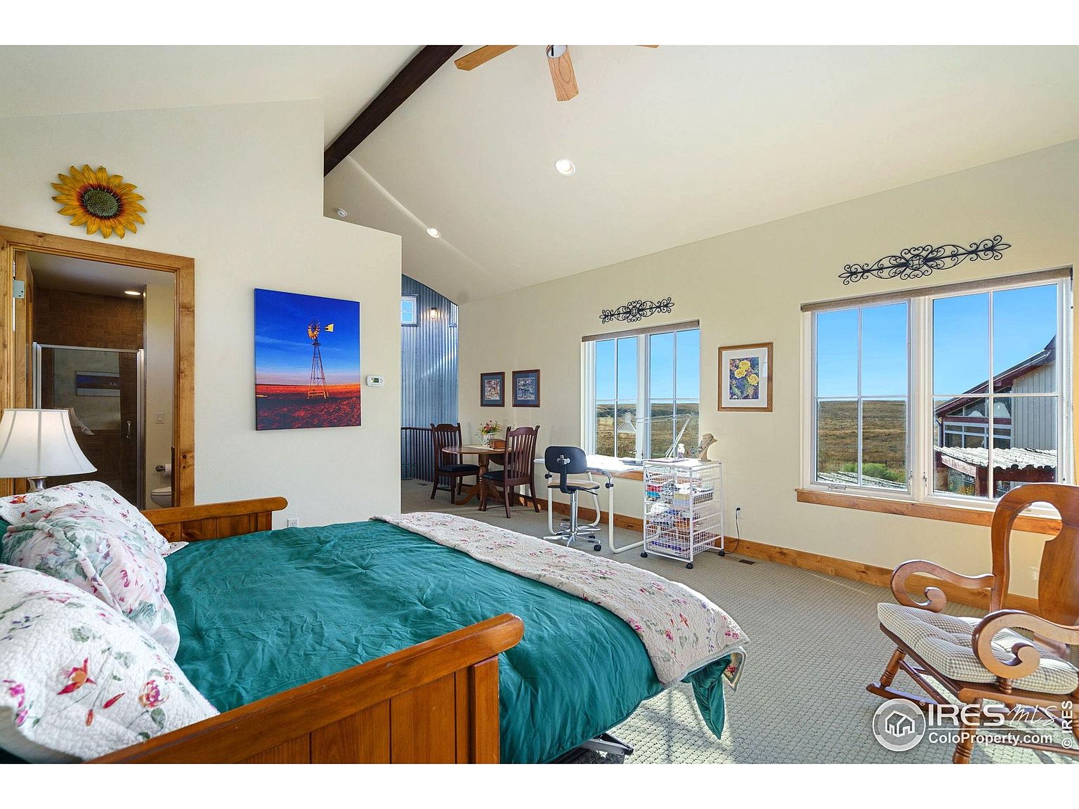This is a primary bedroom featuring a wooden bed with a green comforter and floral pillows. The room has two large windows offering natural light and views of the landscape. There is also a small dining area and a desk with a rolling cart, suggesting a multi-functional space.