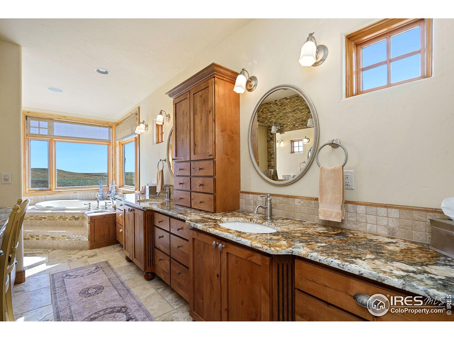 This is a well-appointed primary bathroom featuring a double vanity with granite countertops and wooden cabinetry. An oval mirror hangs above one sink, while a window provides natural light. A soaking tub is visible in the background, suggesting a luxurious and relaxing space.