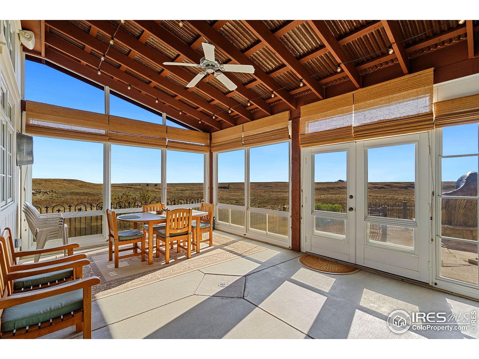 This sunroom features a dining area with a round table and wooden chairs, surrounded by large windows offering expansive views of the landscape. The room has a high, exposed-beam ceiling with a ceiling fan and string lights, creating a warm and inviting atmosphere. The flooring is concrete with geometric patterns, and the windows are covered with woven blinds.