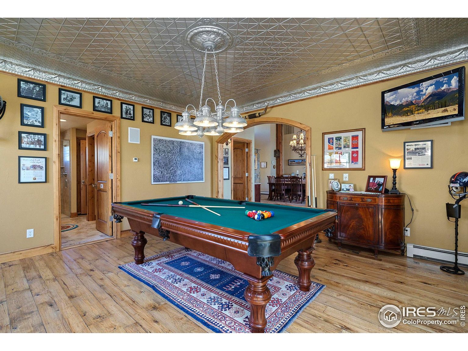 This interior shot showcases a well-appointed living room featuring a billiard table as a central element. The room is decorated with framed pictures, a decorative ceiling, and a wooden cabinet, creating a warm and inviting atmosphere. A television is mounted on the wall, and the room opens to a dining area, suggesting a spacious and comfortable living space.