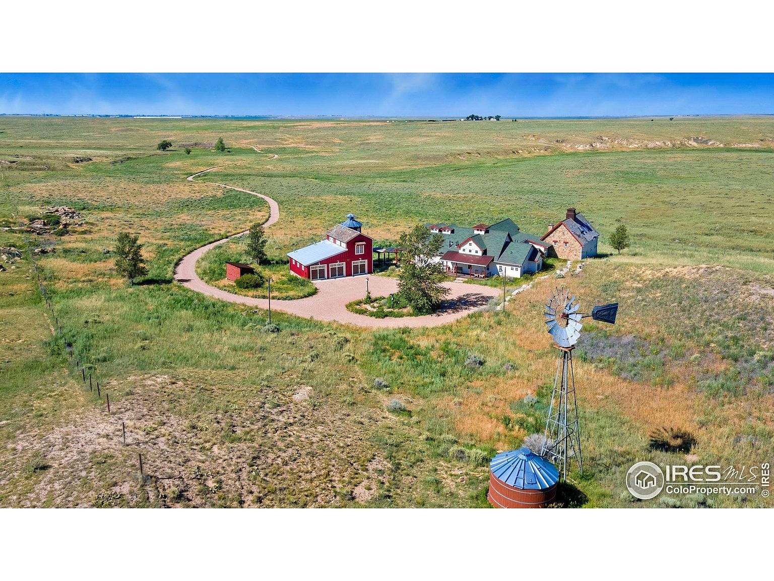 This aerial shot showcases a sprawling property featuring a main house with a green roof, a red barn, and a stone building, all connected by a circular driveway. A windmill stands prominently in the foreground, adding a rustic touch. The landscape is a mix of grassy fields and open plains, creating a sense of vastness and seclusion.