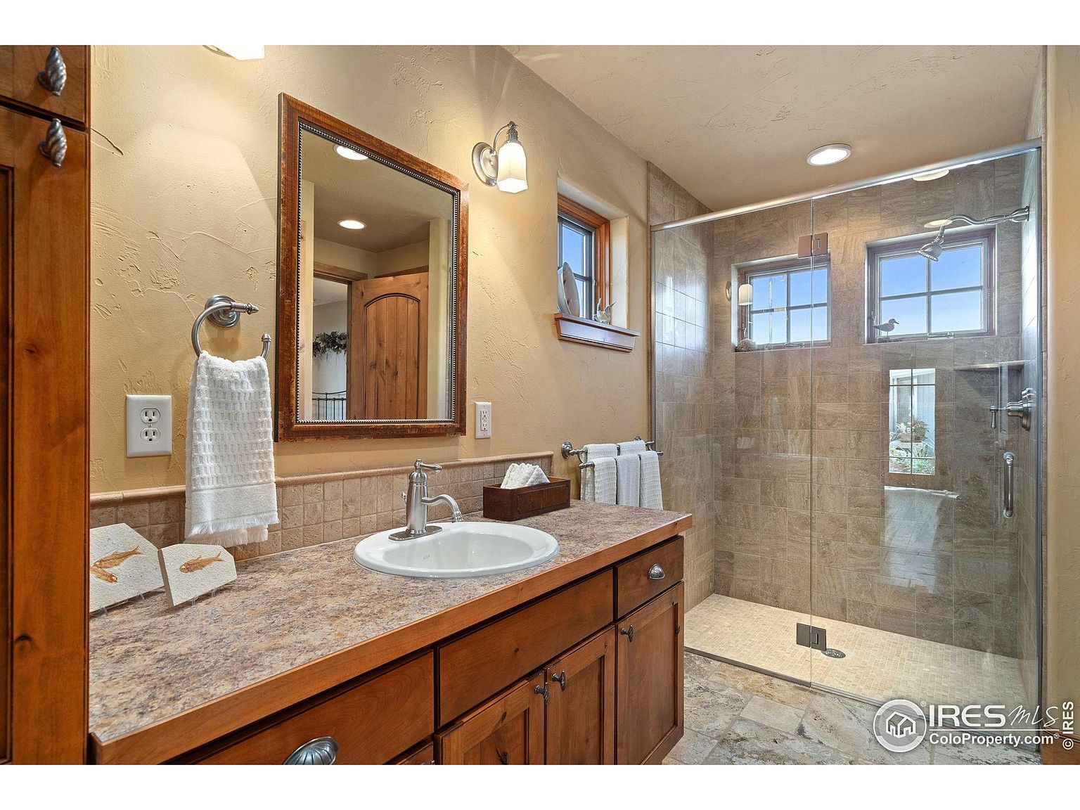 This is a well-lit primary bathroom featuring a wooden vanity with a light-colored countertop and a white sink. A framed mirror hangs above the sink, and a glass-enclosed shower with tiled walls and windows is visible to the right. The overall style is rustic and inviting, with warm tones and natural materials.