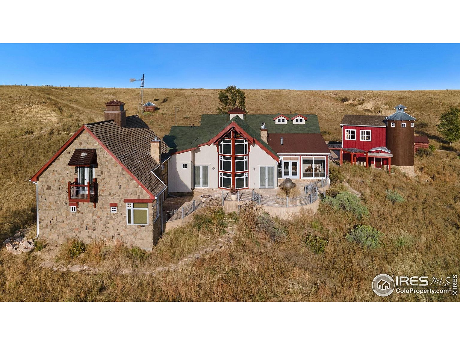 This aerial view showcases a unique multi-building estate nestled on a grassy hillside under a clear blue sky. The property features a stone-clad house with a balcony, a modern structure with large windows, and a red barn-like building with an attached silo. The landscape is characterized by rolling hills and natural vegetation, creating a serene and private setting.