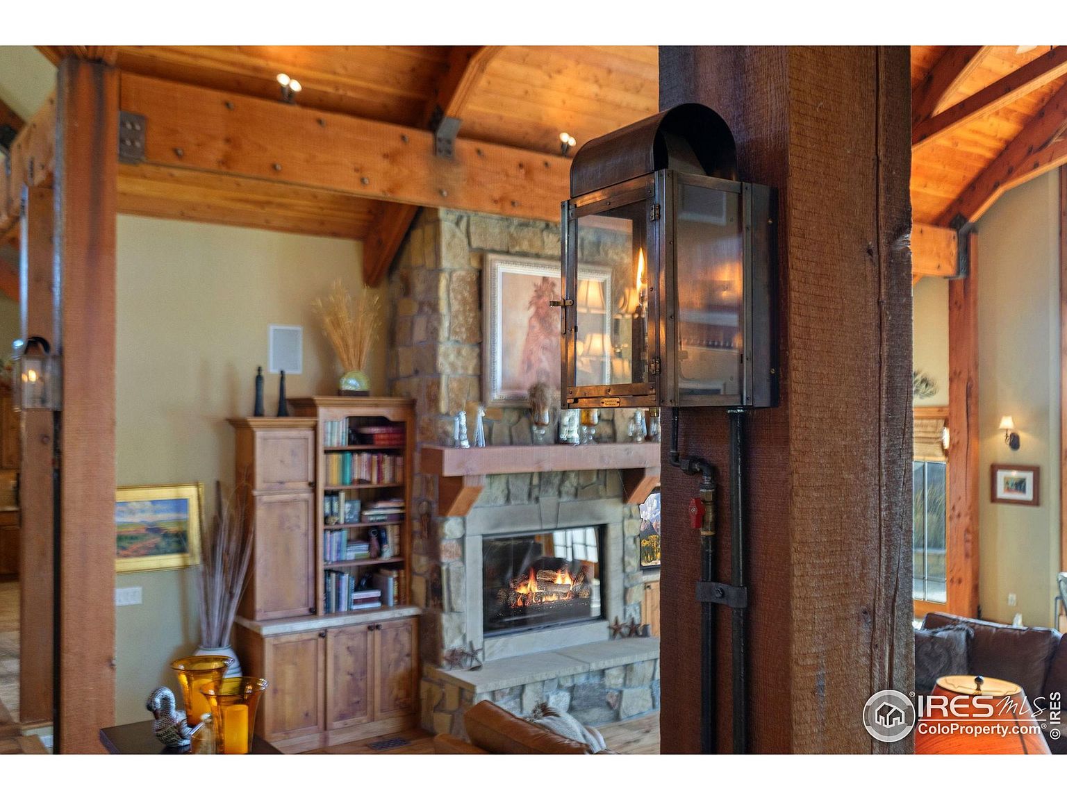 This inviting living room features exposed wooden beams and a stone fireplace, creating a warm and rustic atmosphere. A built-in bookcase adds functionality and charm, while a unique lantern-style light fixture mounted on a wooden post provides ambient lighting. The room exudes a cozy and comfortable vibe, perfect for relaxation and gatherings.