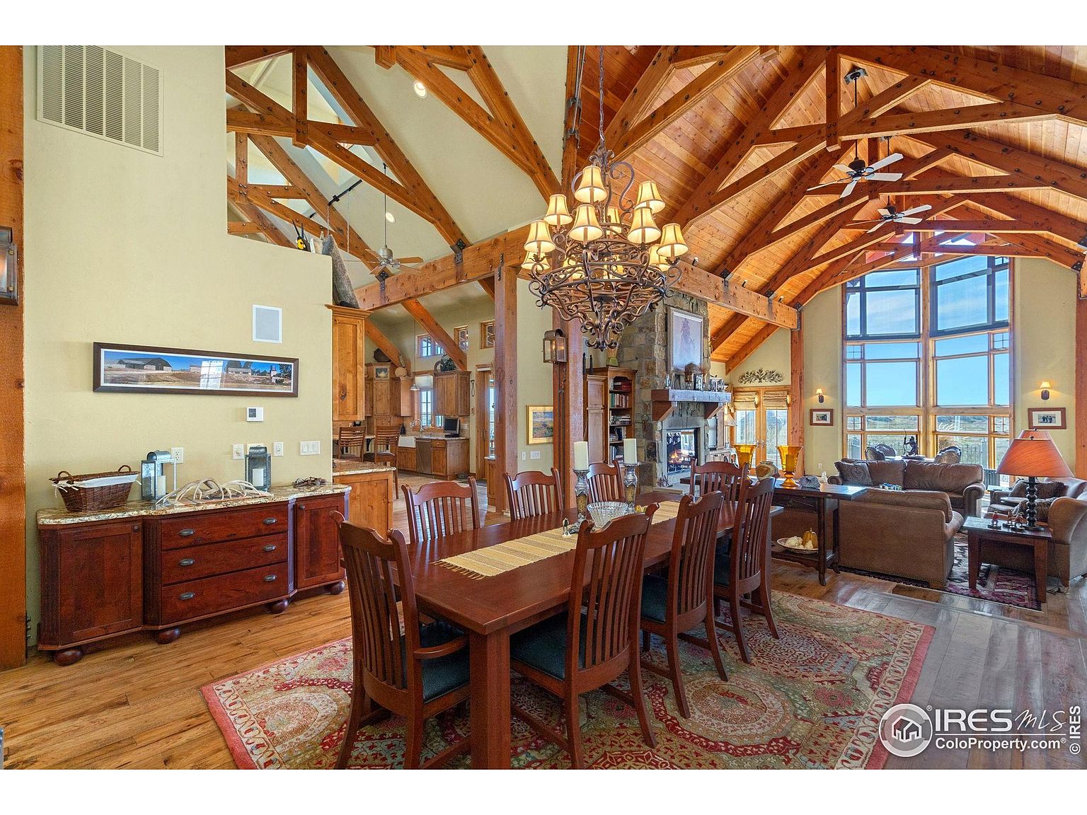 This is an interior shot of a dining room featuring a large wooden dining table with chairs, set on an ornate rug. The room boasts a high, vaulted ceiling with exposed wooden beams and a large chandelier hanging above the table. A stone fireplace and a glimpse of the living room with comfortable seating are visible in the background, creating a warm and inviting atmosphere.