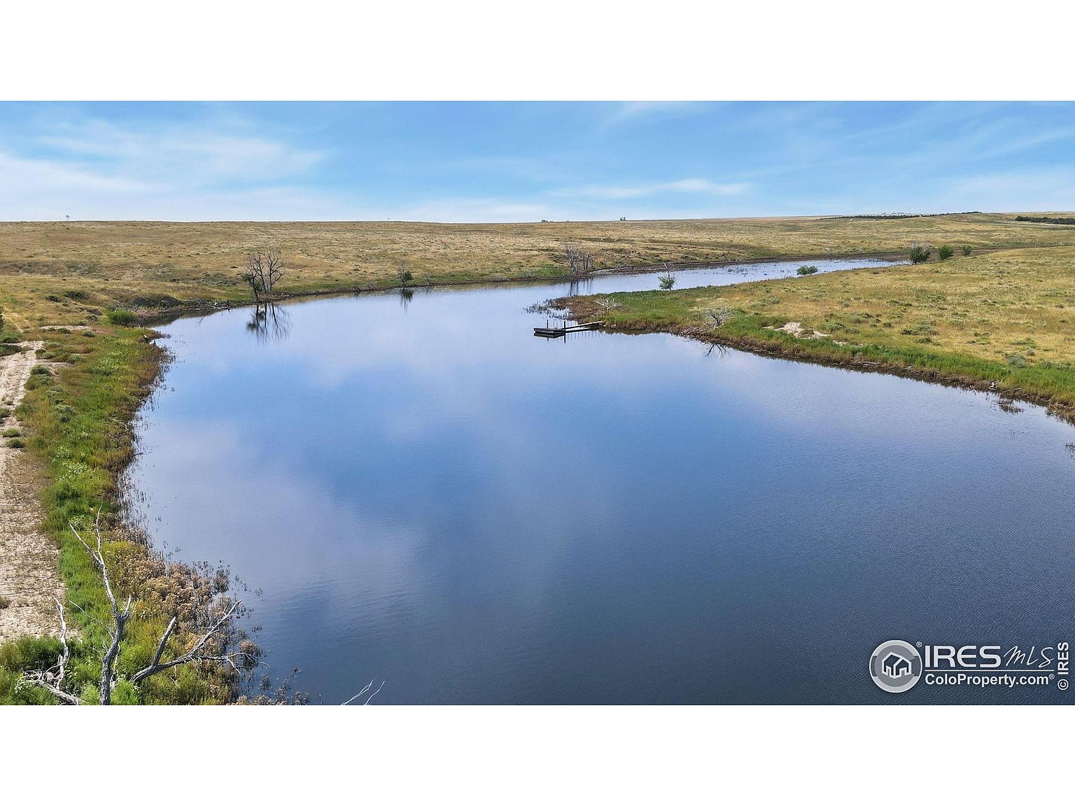 This aerial view showcases a serene pond surrounded by expansive grasslands under a clear blue sky. A small dock extends into the water, suggesting recreational opportunities. The landscape is open and natural, offering a sense of tranquility and seclusion, ideal for a property emphasizing privacy and outdoor living.