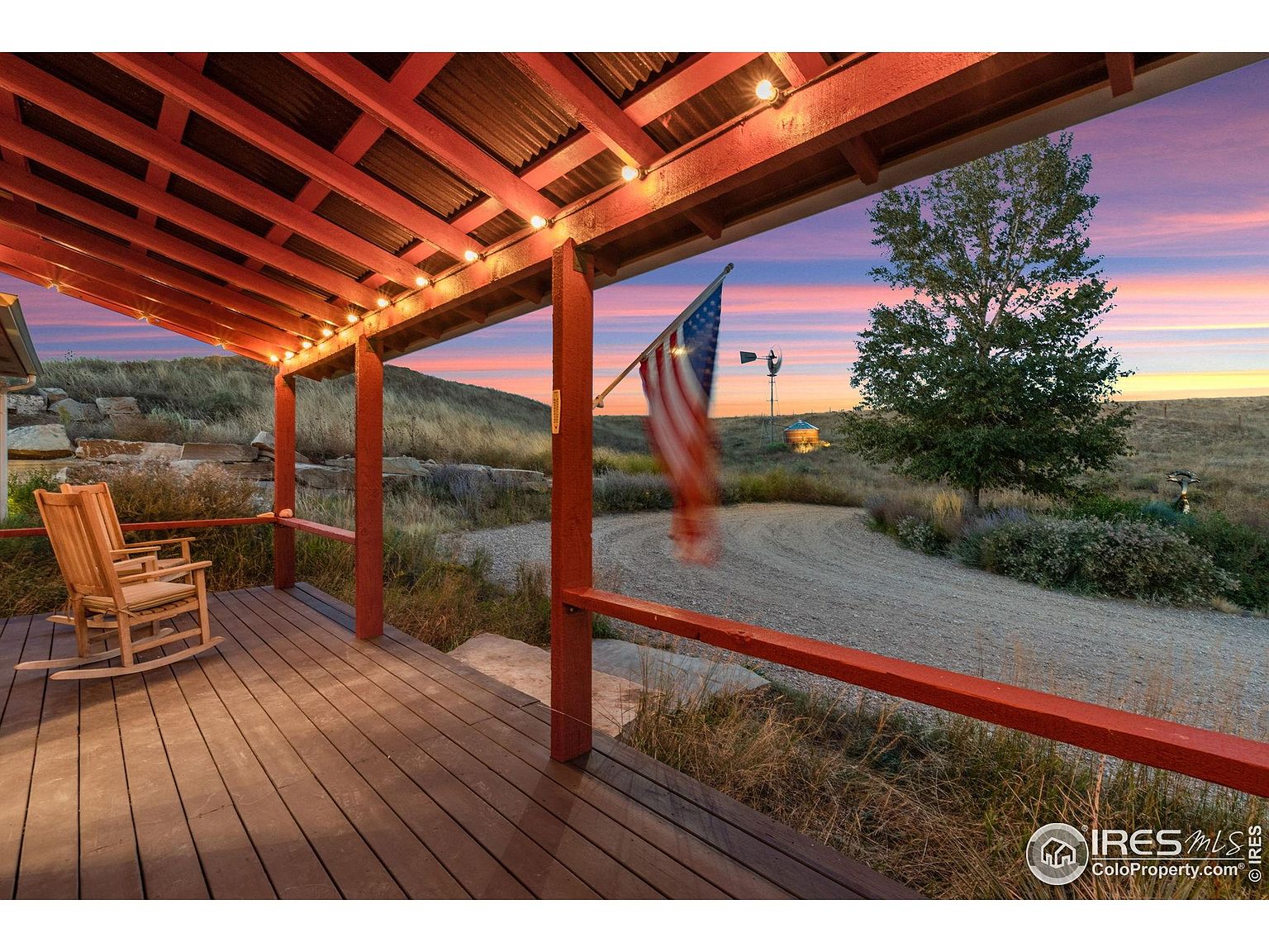 This image showcases a charming patio or deck area with a rustic aesthetic. A wooden rocking chair sits invitingly on the deck, which is covered by a wooden roof structure adorned with string lights. In the background, a picturesque landscape unfolds, featuring a windmill, an American flag, and a serene sunset sky, creating a tranquil and inviting outdoor living space.