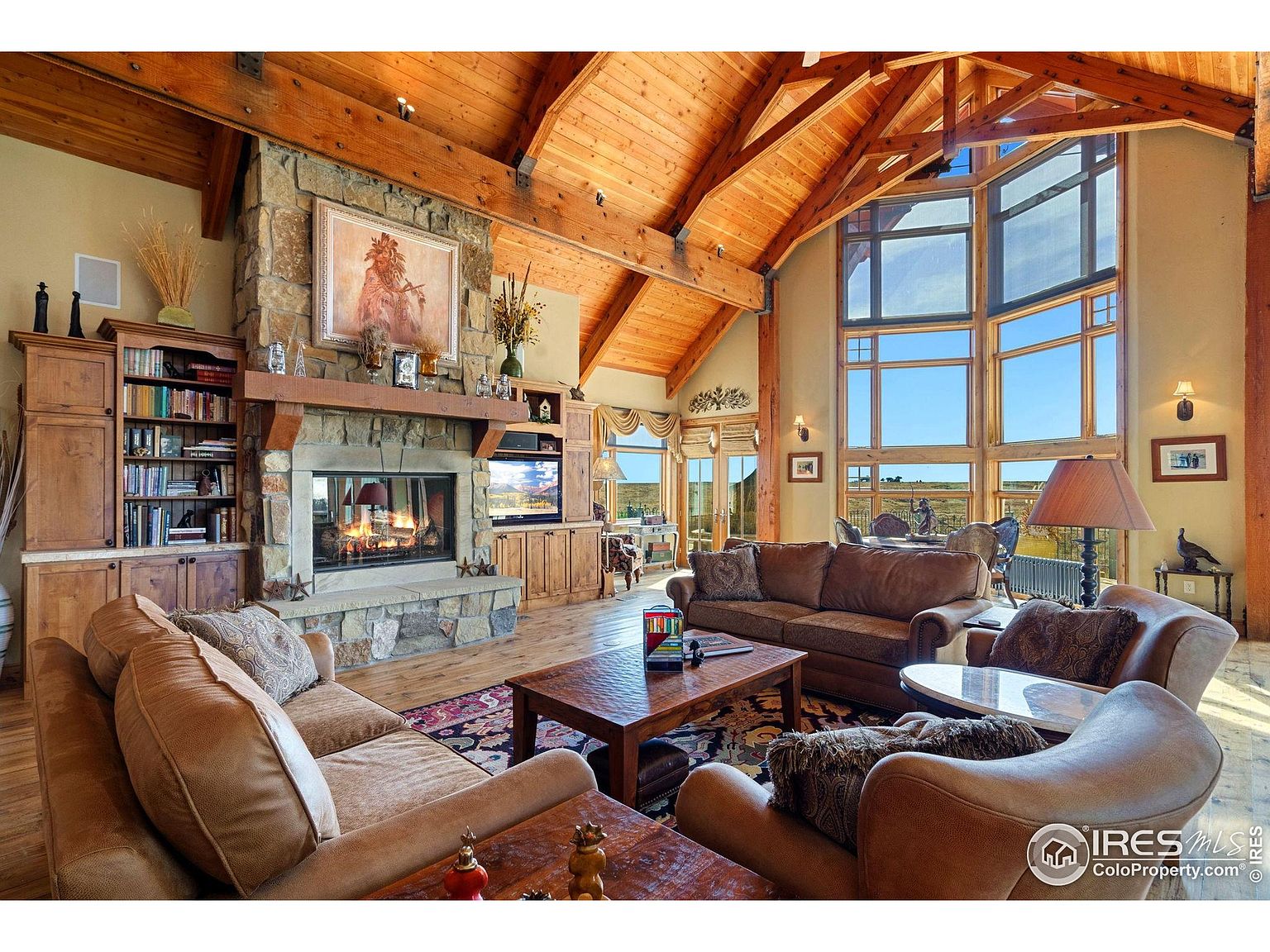 This is a spacious living room featuring a high vaulted ceiling with exposed wooden beams and a large stone fireplace. The room is furnished with comfortable brown leather sofas and armchairs, arranged around a wooden coffee table on a patterned rug. Large windows offer a view of the landscape, creating a warm and inviting atmosphere.
