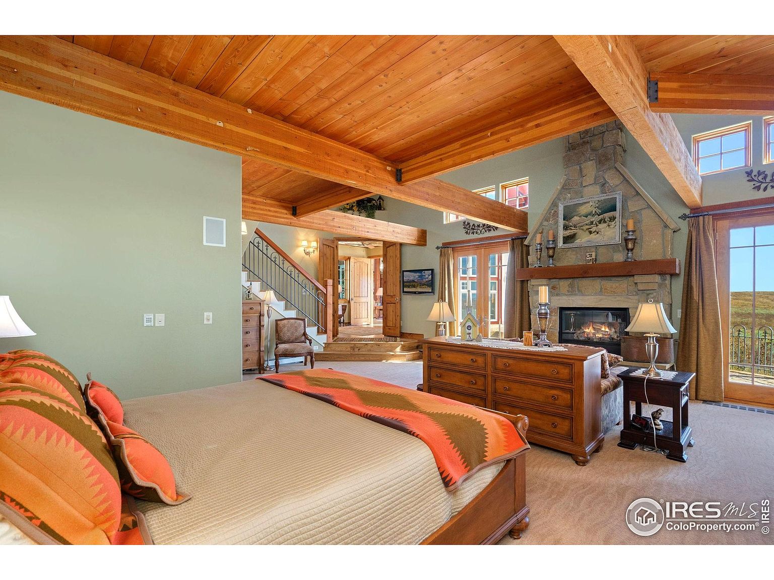 This is a primary bedroom featuring a stone fireplace, wooden beams on the ceiling, and a large bed with patterned bedding. The room has a rustic yet elegant feel, with natural light streaming in from the windows and doors. A dresser and side table provide storage and display space.