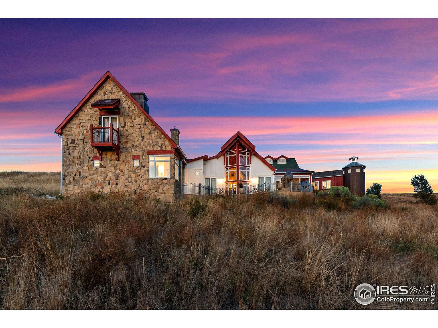This image showcases the front view of a unique and charming estate at sunset. The property features a stone-clad main house with a balcony, connected to a white building with large windows, and a red barn-like structure with a silo. The landscape is dominated by tall, dry grass, creating a rustic and picturesque setting.