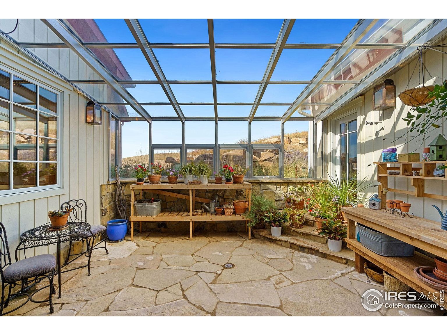 This sunroom features a glass roof and windows, allowing ample natural light to flood the space. The floor is paved with flagstone, and the room is decorated with various potted plants and wooden shelving units. The overall impression is a bright and inviting space for relaxation and gardening.