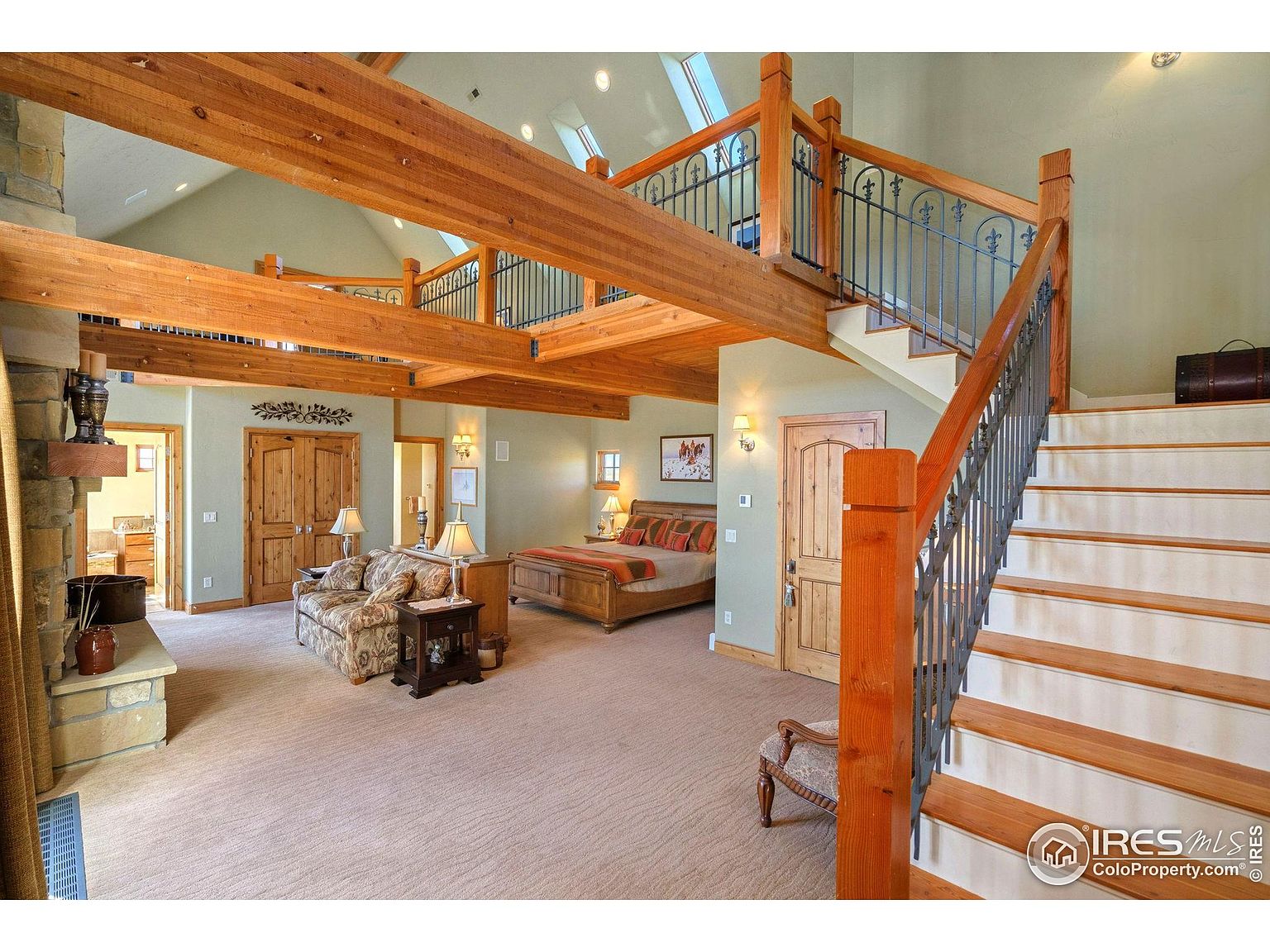 This is a spacious primary bedroom featuring exposed wooden beams and a loft area. The room includes a king-size bed, a seating area with a floral sofa, and a stone fireplace. A staircase leads to the loft, adding architectural interest and suggesting a luxurious, open layout.
