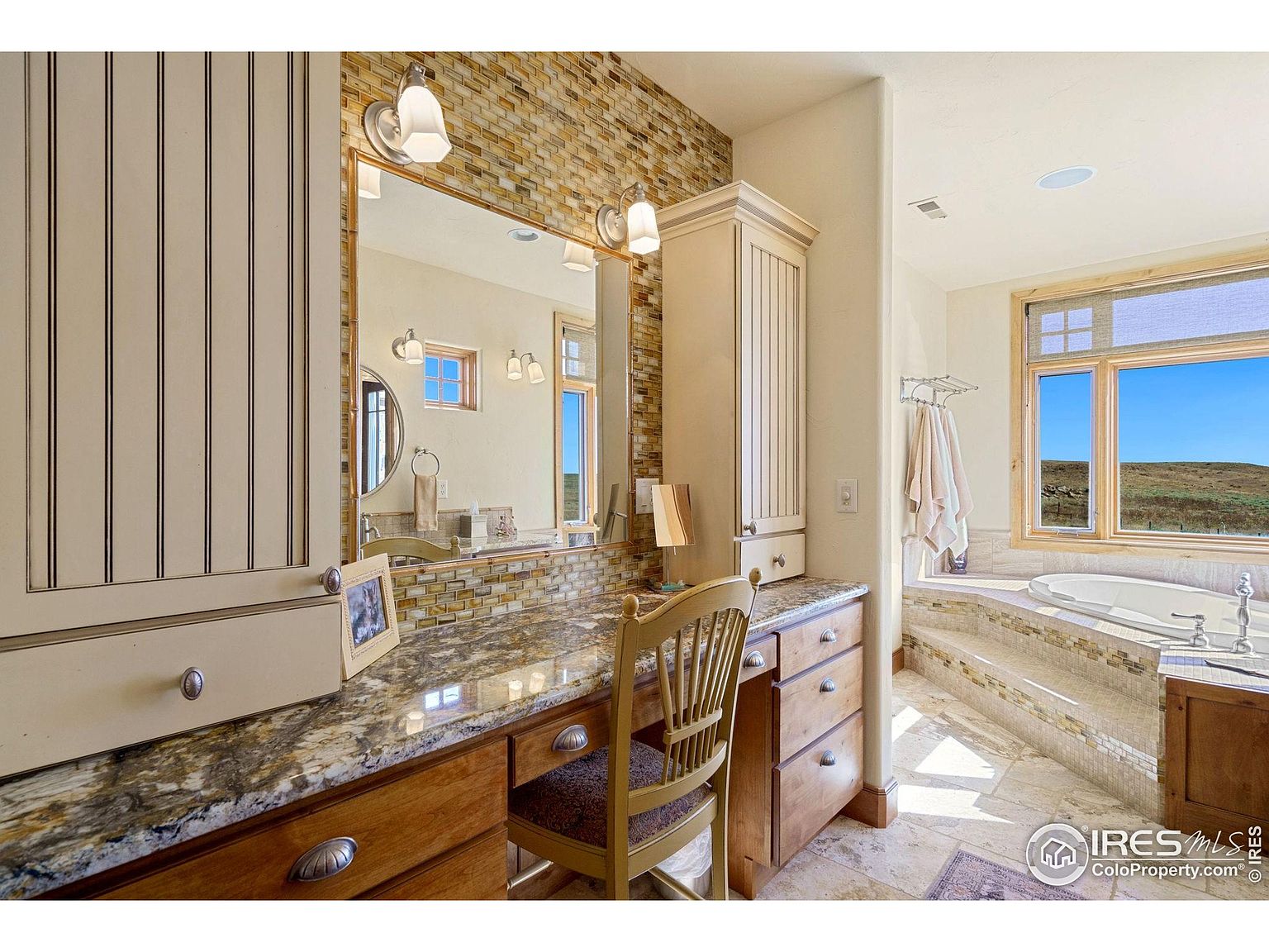 This is a primary bathroom featuring a vanity area with a granite countertop, wooden cabinets, and a large mirror with sconce lighting. The bathroom also includes a soaking tub with a view of the landscape through a large window. The overall style is luxurious and well-lit, creating a relaxing atmosphere.