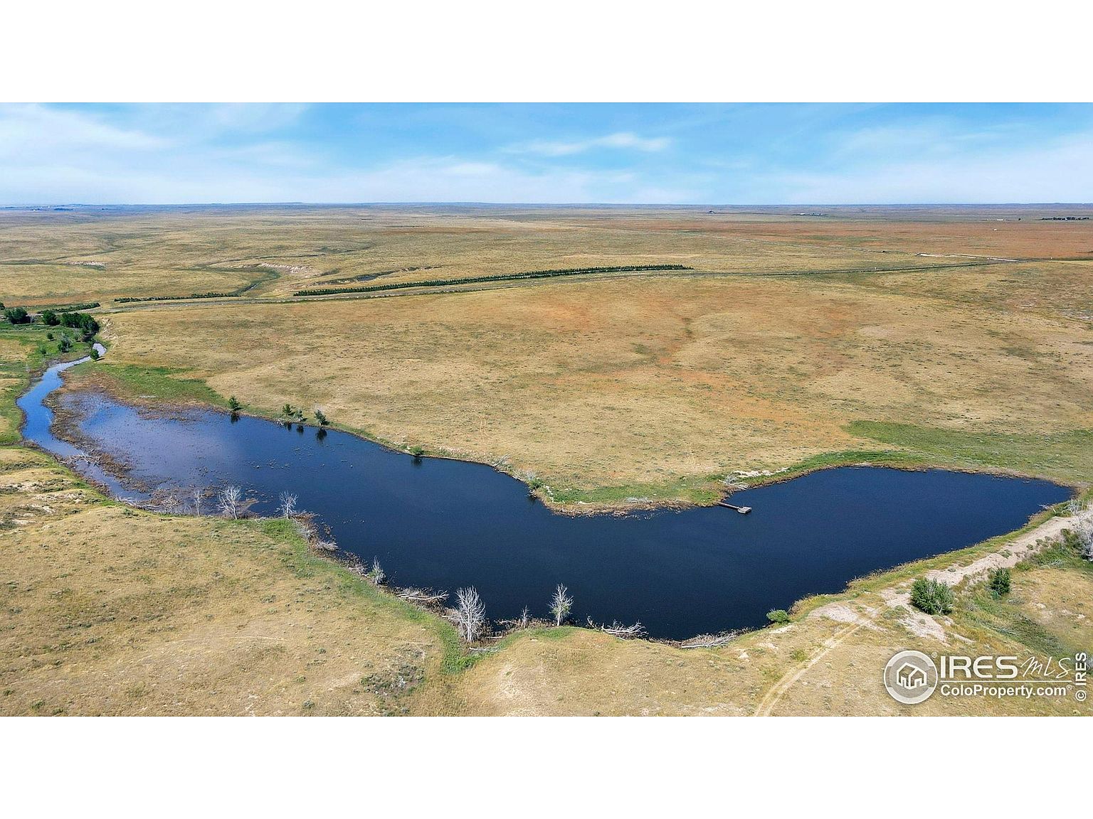 This aerial shot showcases a vast expanse of land featuring a large, irregularly shaped pond or lake. The surrounding terrain is primarily dry grassland, stretching out to the horizon under a partly cloudy sky. The image emphasizes the property's size and natural water feature, highlighting its potential for recreational or agricultural use.