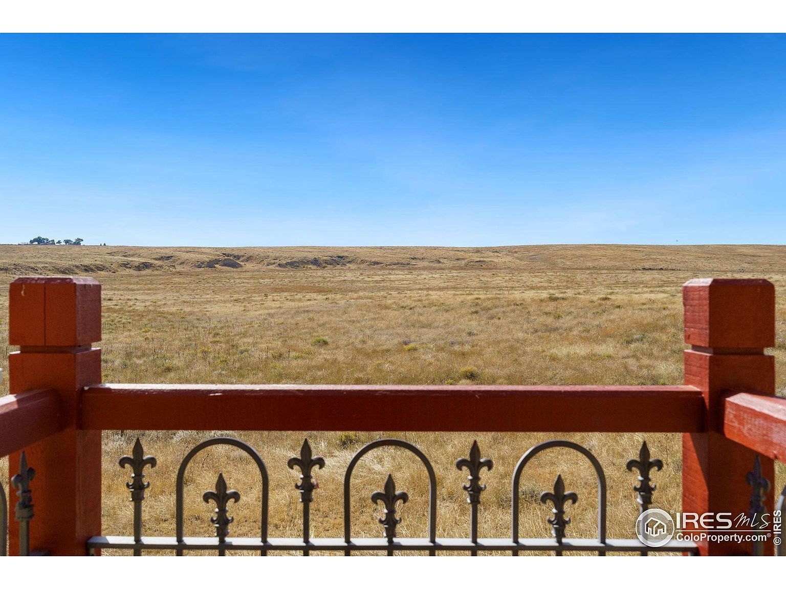 This image showcases a view from a patio or balcony, featuring a decorative railing in the foreground. Beyond the railing, a vast, open field stretches to the horizon under a clear blue sky. The scene evokes a sense of tranquility and expansive outdoor living.