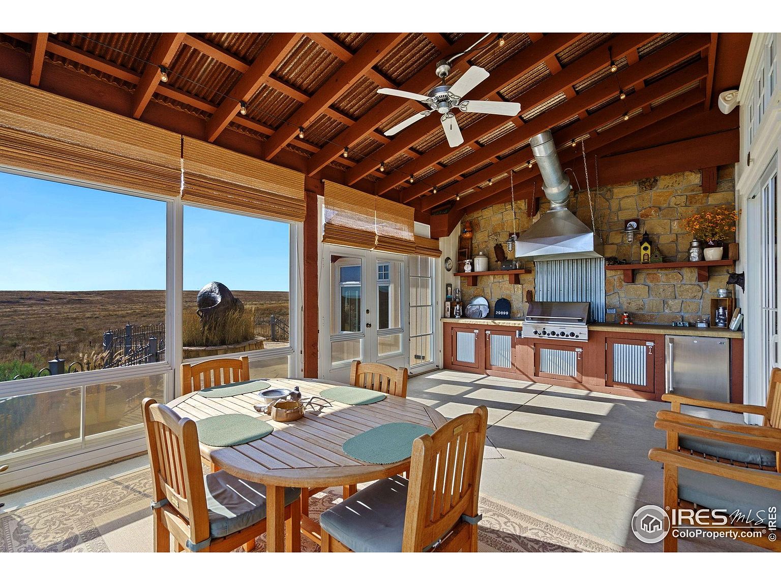 This image showcases a well-appointed outdoor patio or sunroom featuring a built-in kitchen area with stone accents, stainless steel appliances, and wooden cabinetry. A round wooden dining table with chairs sits near large windows offering scenic views of the landscape. The space is covered with a wooden ceiling and features a ceiling fan, creating a comfortable and stylish outdoor living area.