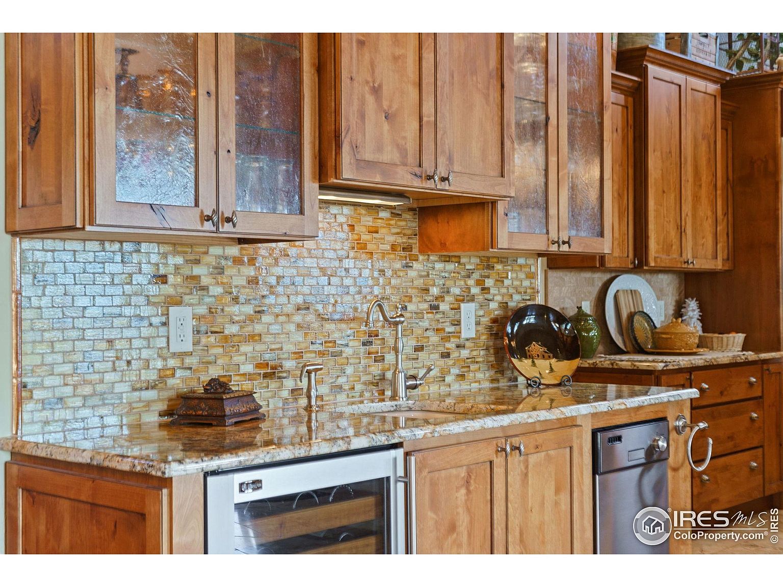 This is a well-appointed kitchen featuring wooden cabinetry, a mosaic tile backsplash, and granite countertops. Stainless steel appliances, including a wine cooler and dishwasher, add a modern touch. The kitchen has a warm and inviting feel, with ample storage and counter space.