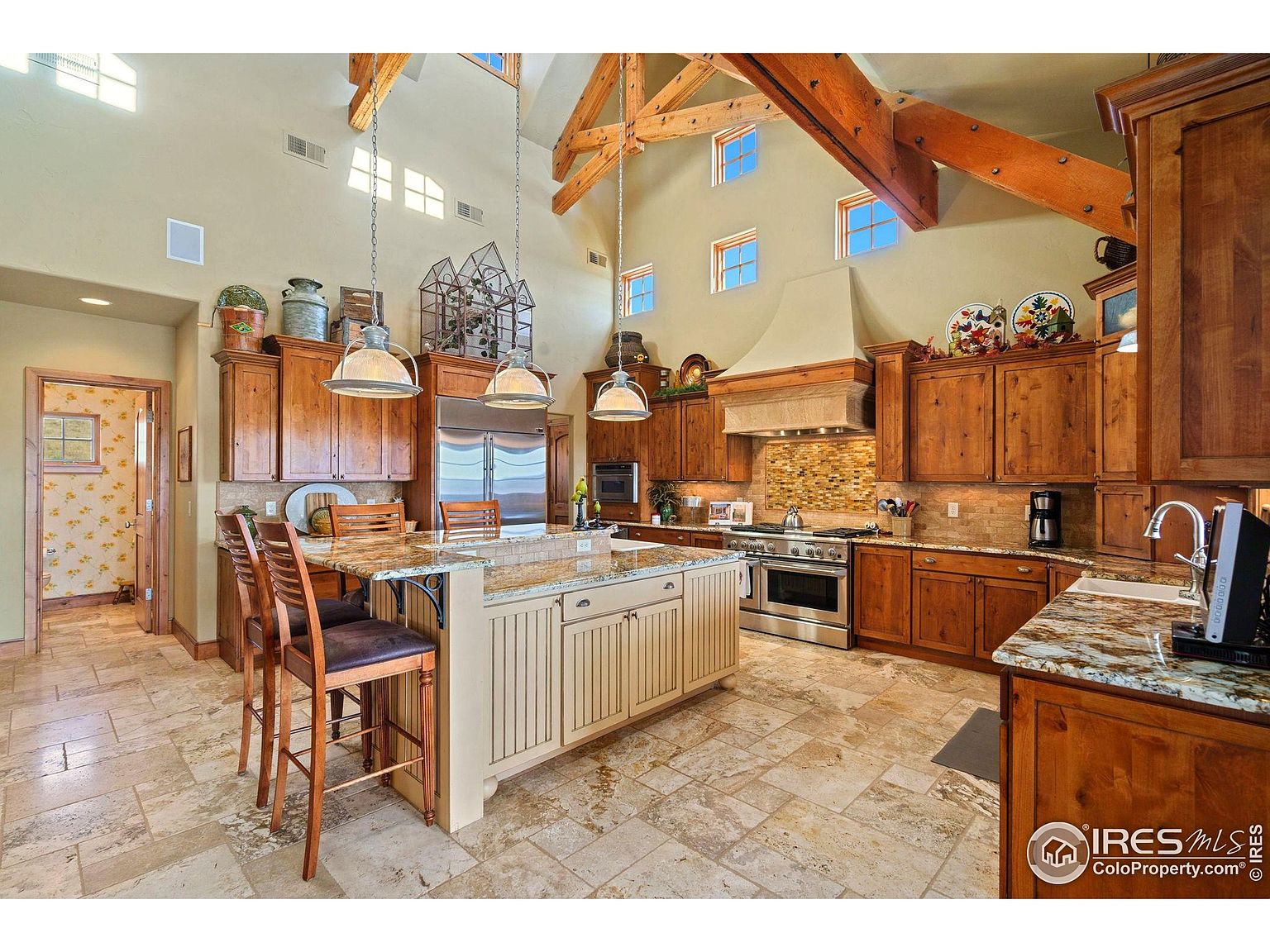 This is a spacious kitchen with high ceilings, exposed wooden beams, and custom cabinetry. The large island features a granite countertop and provides ample seating. Stainless steel appliances and a decorative backsplash add to the kitchen's appeal, creating a warm and inviting atmosphere.