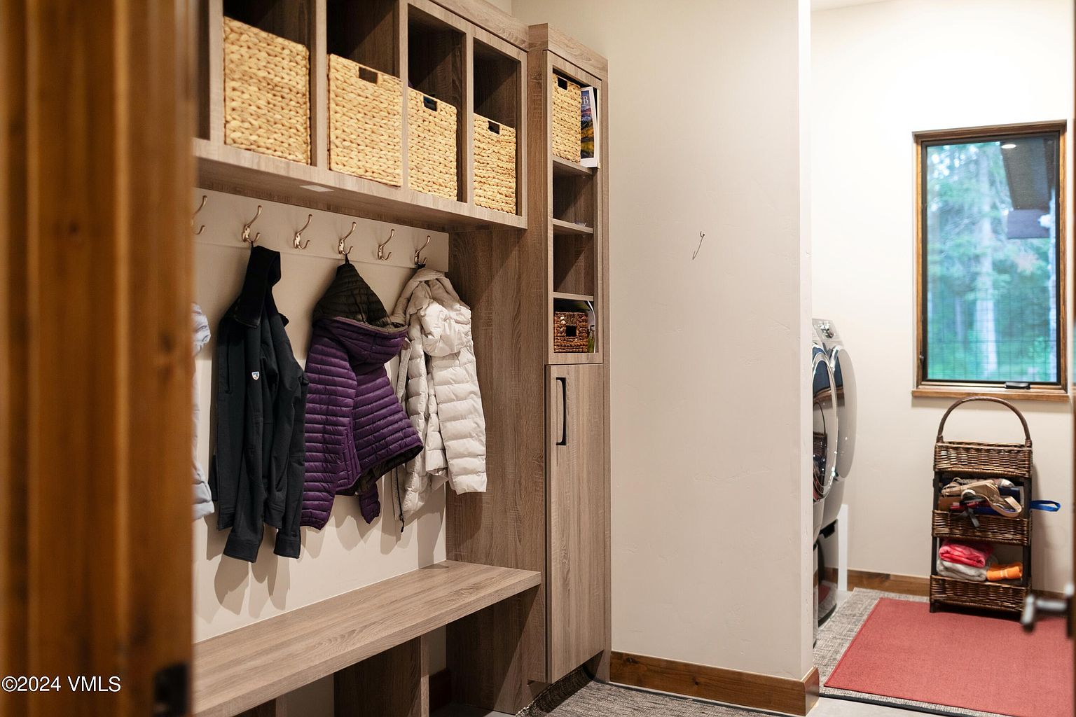 This interior shot showcases a well-organized laundry room with custom built-in storage. The space features a bench, coat hooks, and woven baskets for storage, creating a functional and stylish area. A laundry basket and a glimpse of the washer and dryer are visible, suggesting a practical and efficient design.