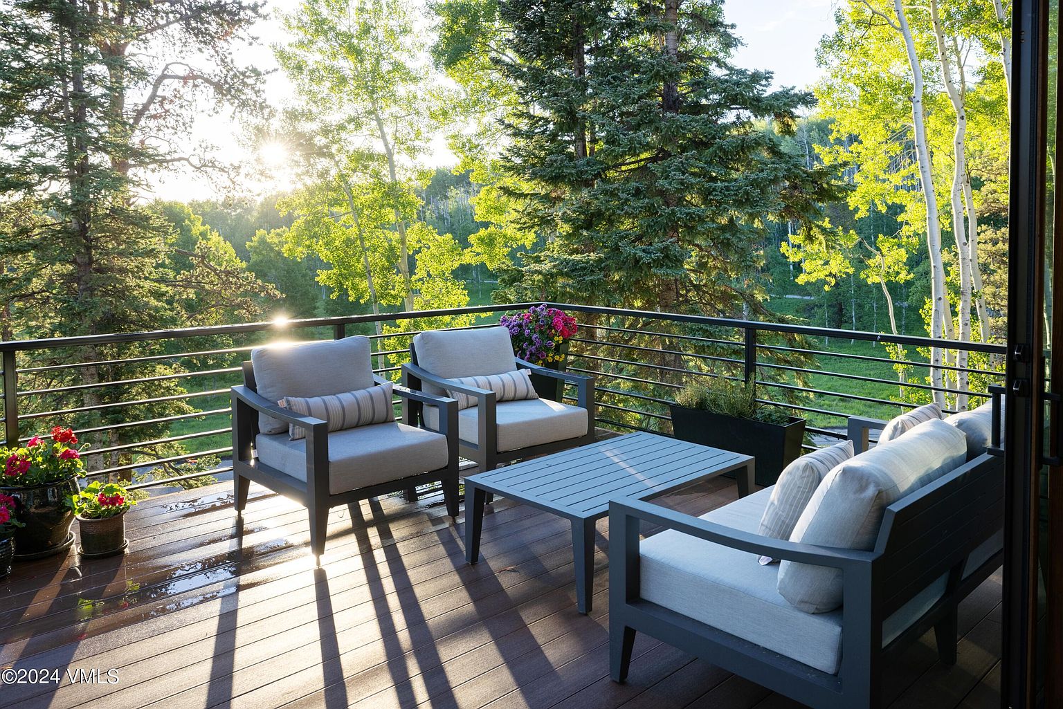 This image showcases a beautifully designed outdoor patio or deck area, perfect for relaxation and entertaining. The space features comfortable seating arrangements, including chairs and a sofa, arranged around a coffee table. The deck is surrounded by lush greenery and trees, offering a serene and private atmosphere, with the sun shining through the trees.