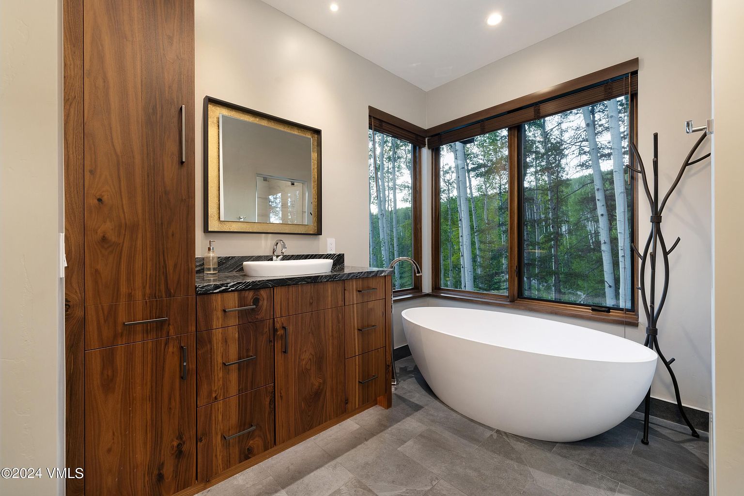 This is a luxurious primary bathroom featuring a modern freestanding bathtub positioned near a large window offering a view of a forest. The vanity area includes a dark countertop, a vessel sink, and a wood-finished cabinet with ample storage. The flooring is a sleek gray tile, and the overall aesthetic is contemporary and serene.