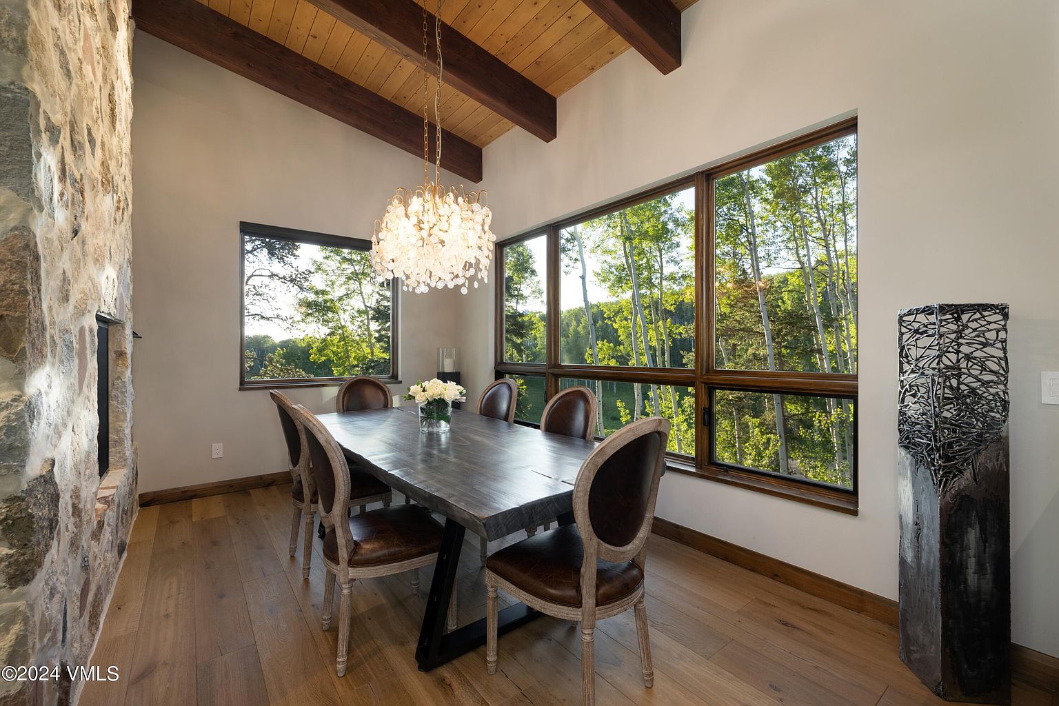 This is an interior shot of a dining room featuring a large wooden table with six chairs, a decorative chandelier, and two windows offering views of the surrounding trees. The room has a rustic yet elegant style, with wooden beams on the ceiling and a stone accent wall, creating a warm and inviting atmosphere perfect for family gatherings or formal dinners.