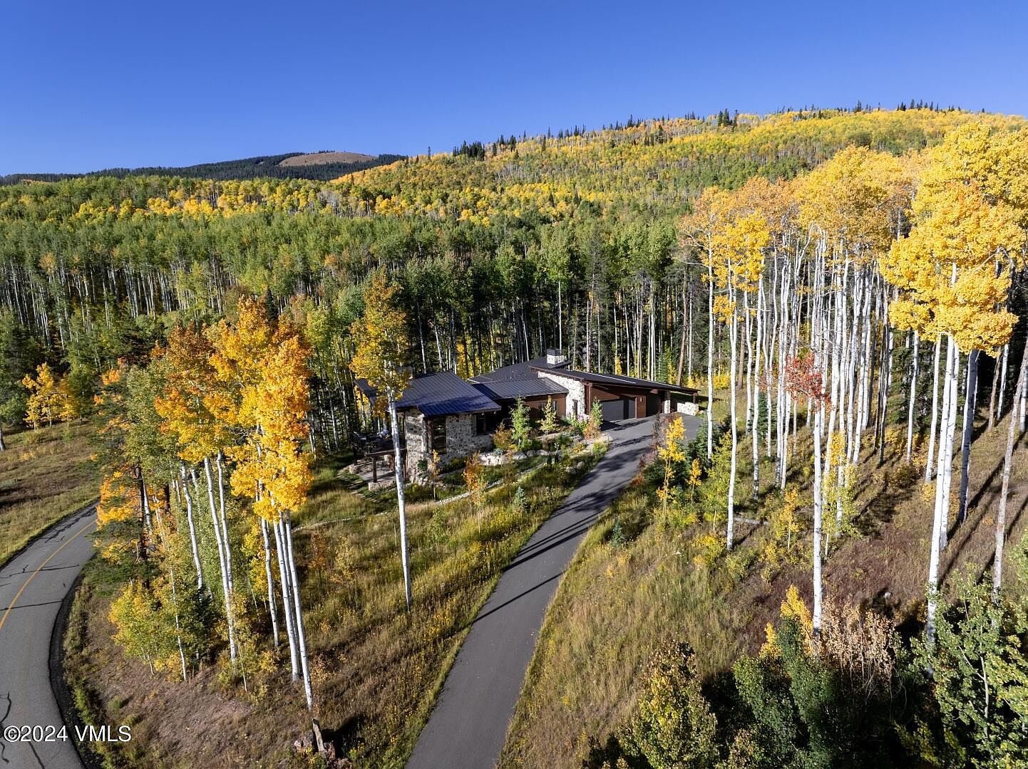 This aerial view showcases a beautiful home nestled among vibrant fall foliage. The house features a stone facade and a dark roof, complemented by a winding driveway. The surrounding landscape is a mix of evergreen and deciduous trees, creating a picturesque and private setting.
