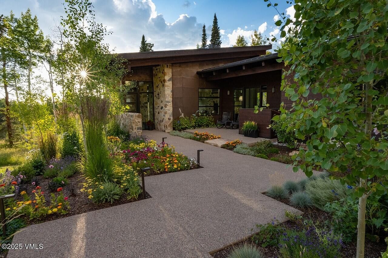 This image showcases the beautifully landscaped yard and garden of a modern home. A paved walkway leads to the entrance, flanked by vibrant flower beds and lush greenery. The architecture features a blend of stone and wood, creating a warm and inviting atmosphere.