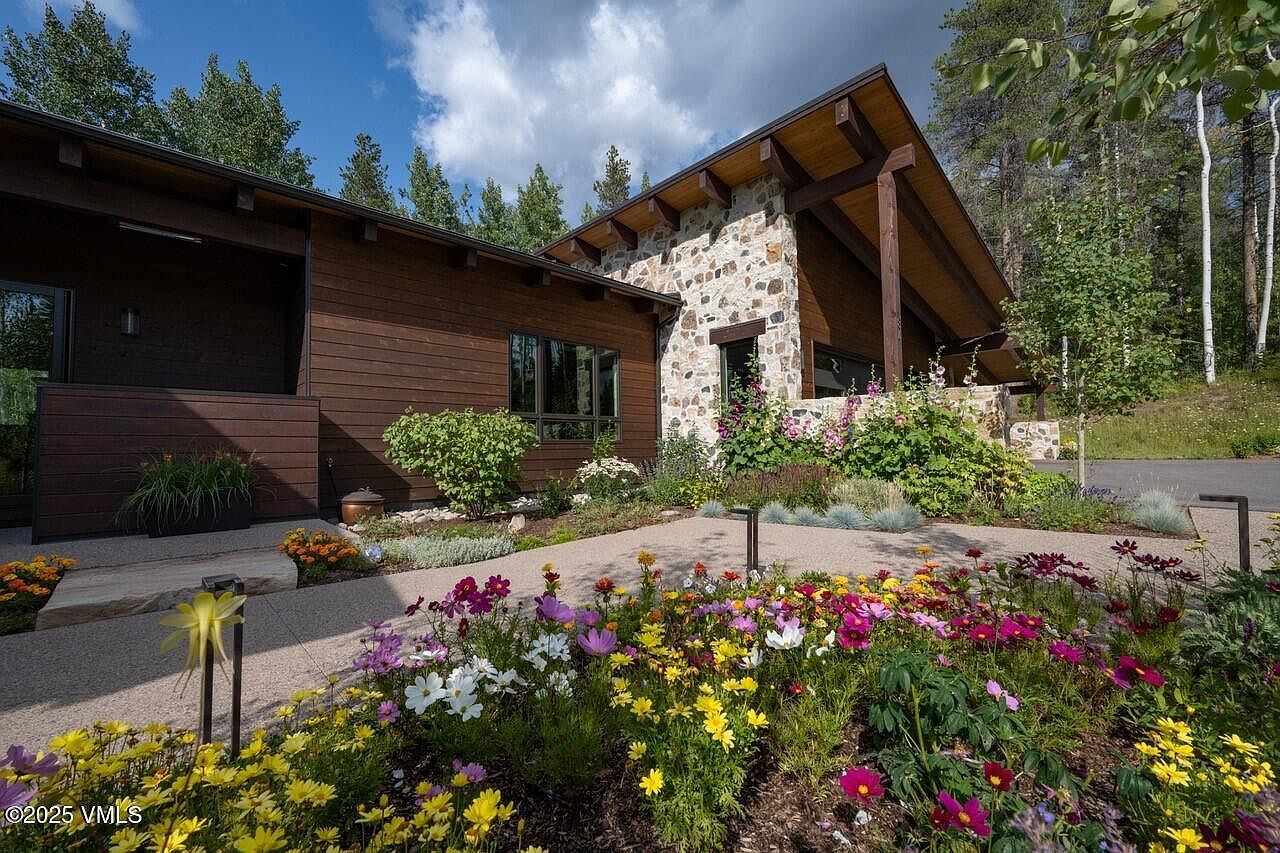 This image showcases the front yard and garden of a modern home. The house features a combination of wood siding and stone accents, with a well-maintained garden filled with colorful flowers in the foreground. A paved walkway leads to the entrance, creating an inviting and aesthetically pleasing curb appeal.
