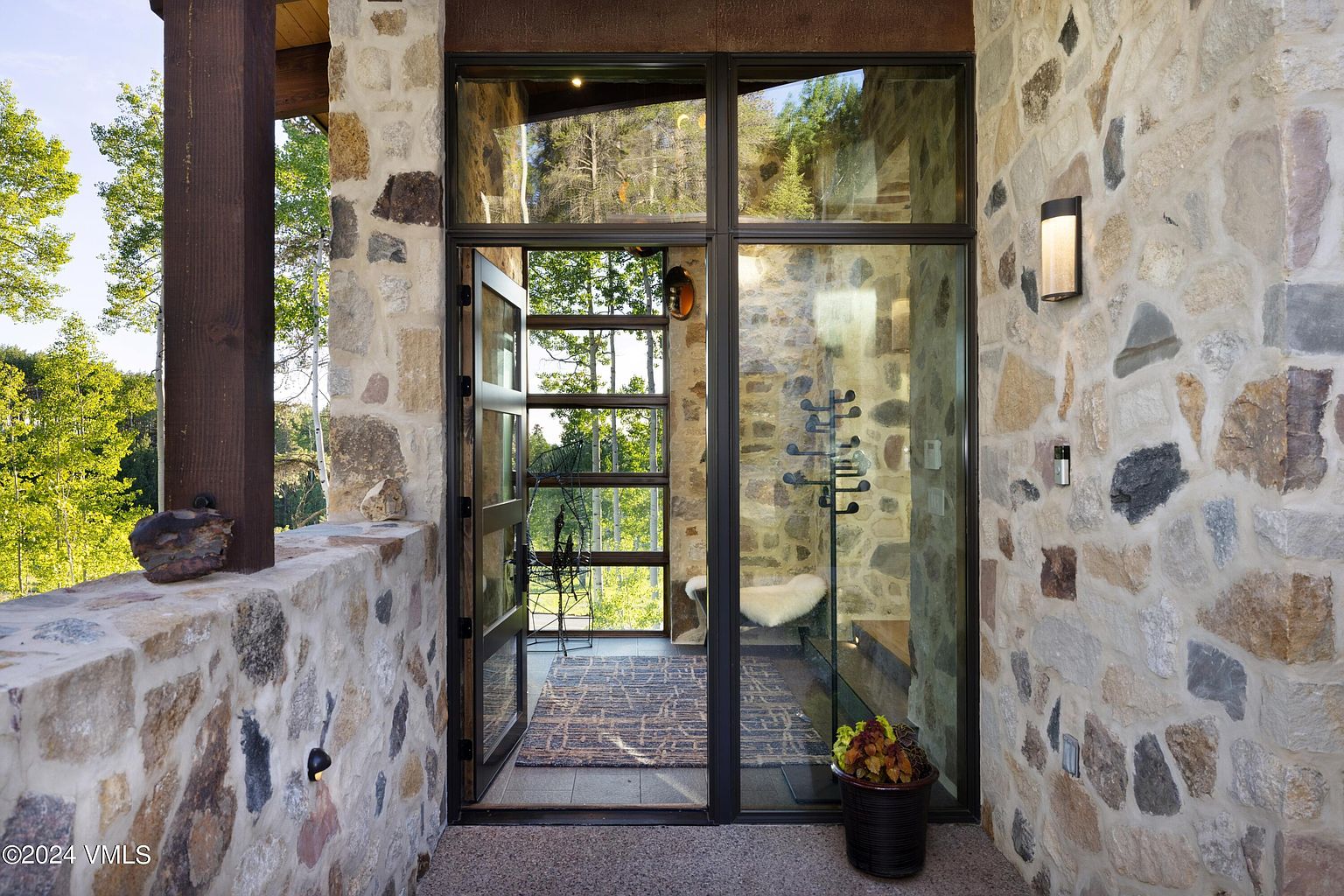 This image showcases a grand entryway featuring a modern glass and metal door framed by rustic stone walls. The open door reveals a glimpse of the interior, including a rug and seating area, while a potted plant adds a touch of greenery. The combination of natural stone and contemporary design creates a sophisticated and inviting entrance.