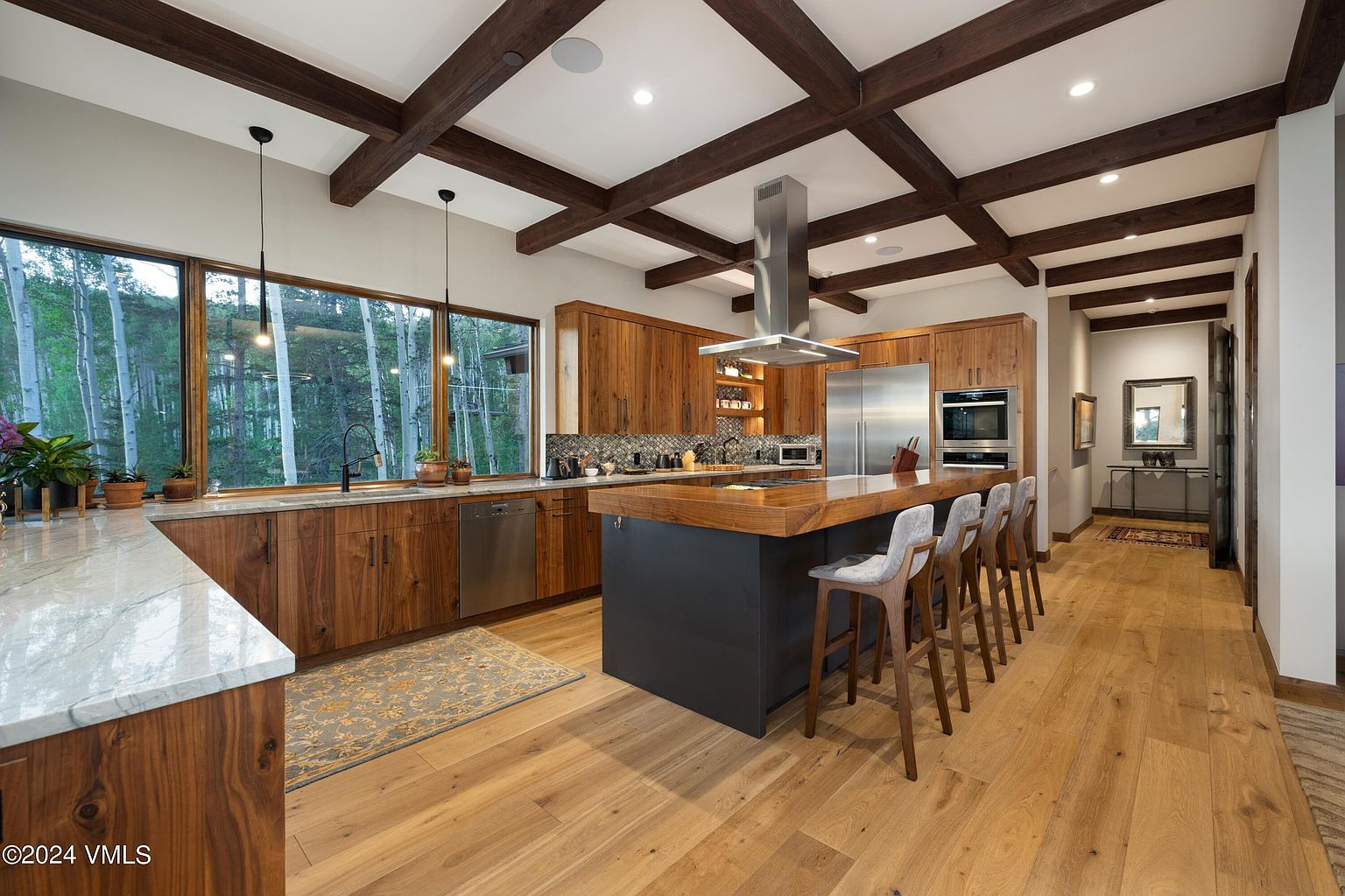 This is a well-lit, modern kitchen featuring wooden cabinetry and a large island with bar seating. The kitchen boasts stainless steel appliances and a unique ceiling design with exposed wooden beams. A large window provides natural light and a view of the surrounding trees, enhancing the kitchen's warm and inviting atmosphere.