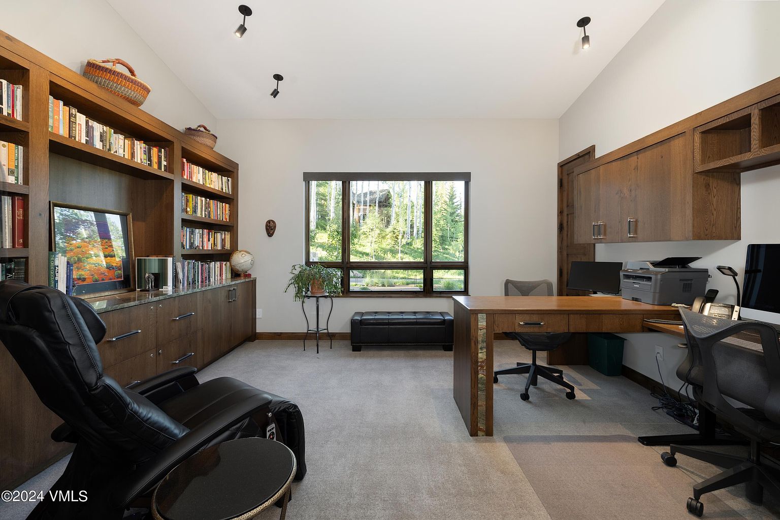 This is an interior shot of a well-appointed home office. The room features custom built-in bookshelves and cabinetry, a large window offering natural light and a view of the outdoors, and a substantial wooden desk with ample workspace. A comfortable leather chair and additional seating create an inviting and functional environment.