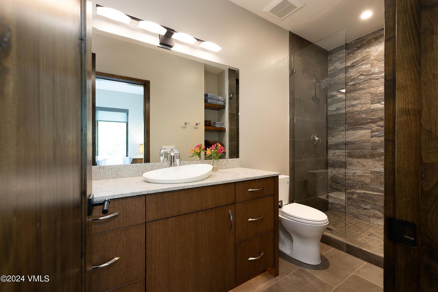 This is a well-lit primary bathroom featuring a modern vanity with a white vessel sink and granite countertop. A large mirror reflects the space, including a built-in shelf with neatly stacked towels. The bathroom also includes a glass-enclosed shower with tiled walls and a toilet, creating a clean and functional space.